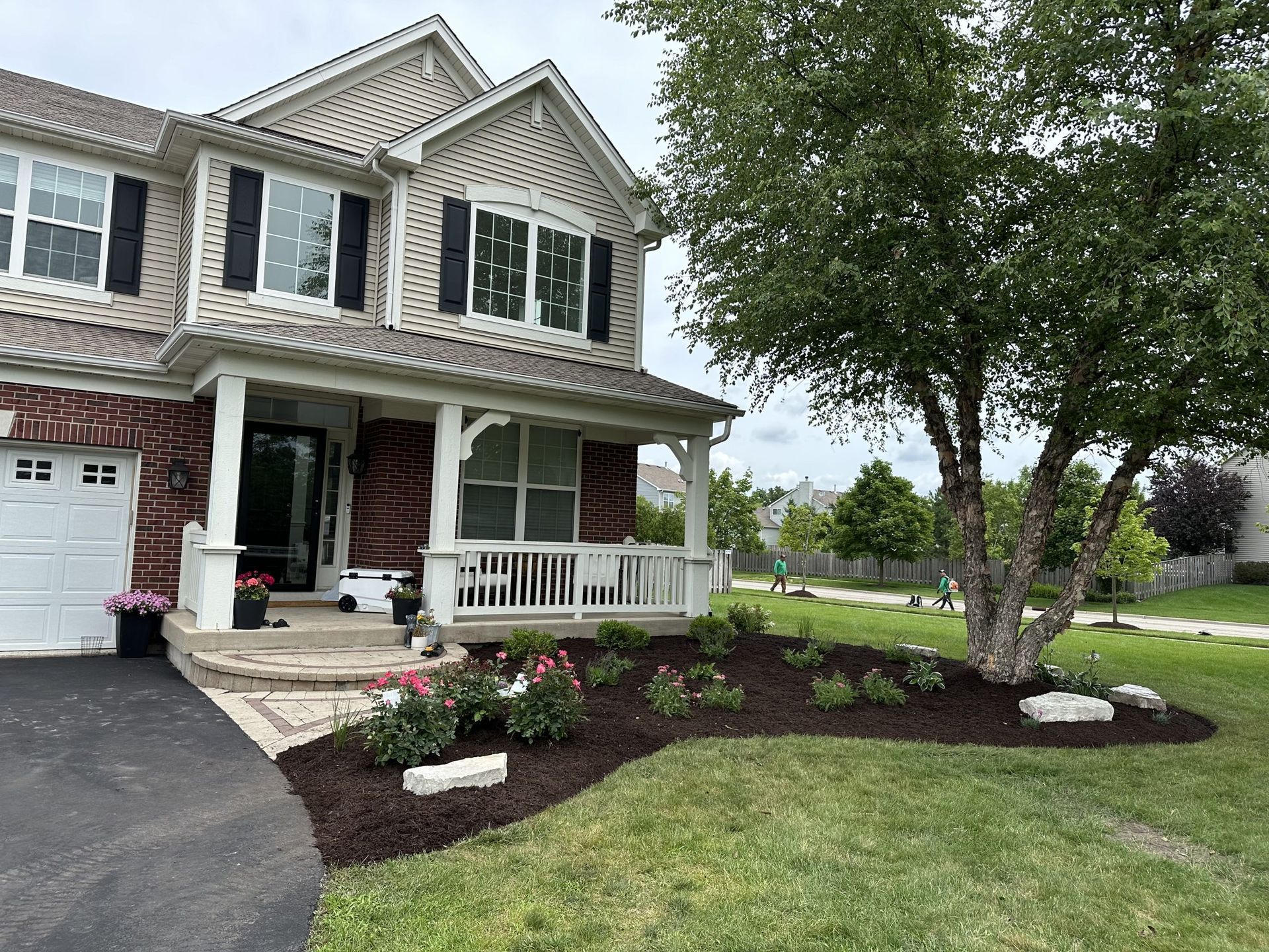 Two-story house with front porch and manicured landscaping, including a tree and flower beds with dark mulch.