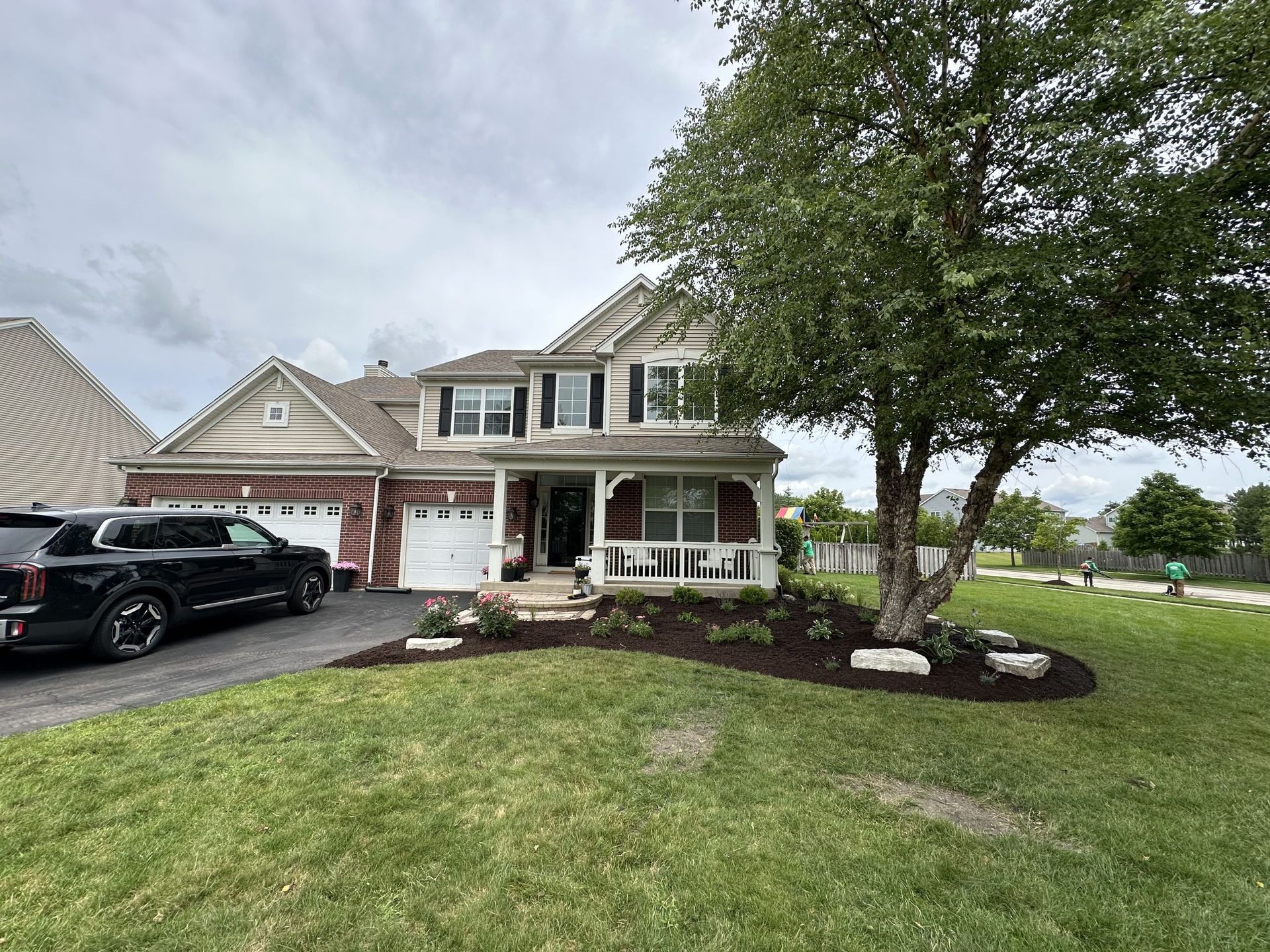 Two-story house with black truck in driveway, landscaping with mulch and tree, green lawn, cloudy sky.