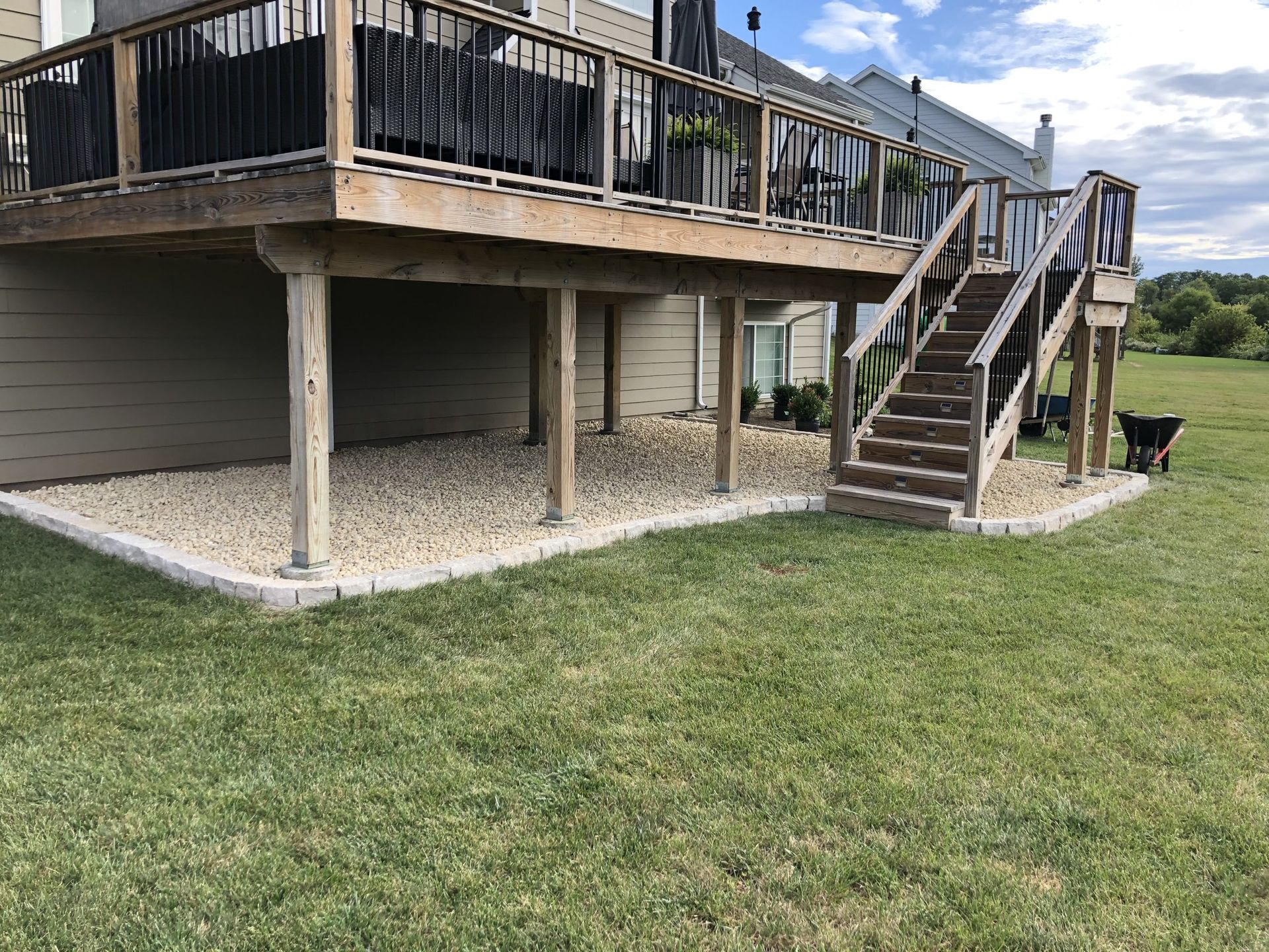 Wooden deck over a gravel bed, accessible via stairs. Located in a yard with green grass and a wheelbarrow.