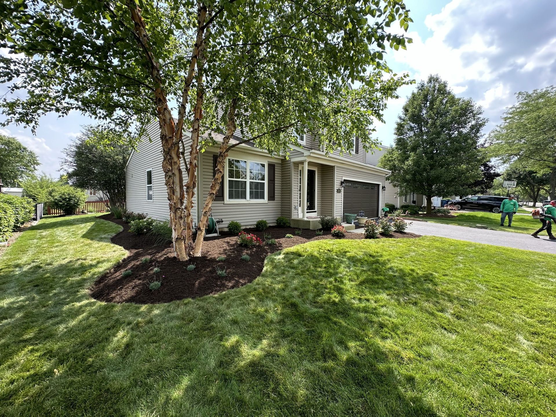 A light-colored house with a landscaped yard and a tree in front. Two people in green are near the driveway.