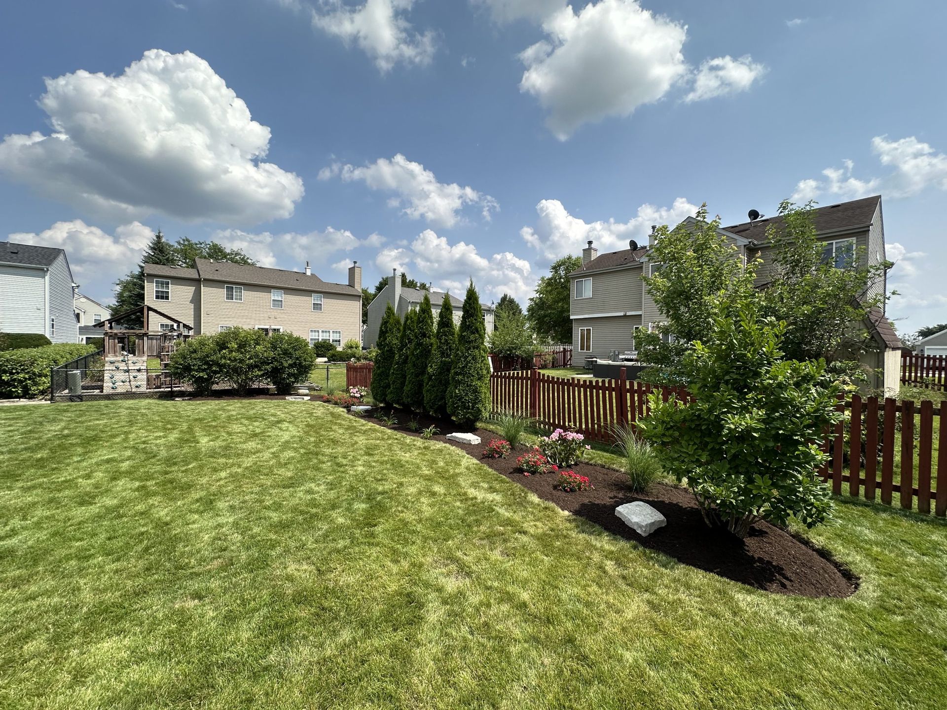 Lush backyard with green grass, brown mulch, and a red picket fence, under a blue sky with fluffy clouds.