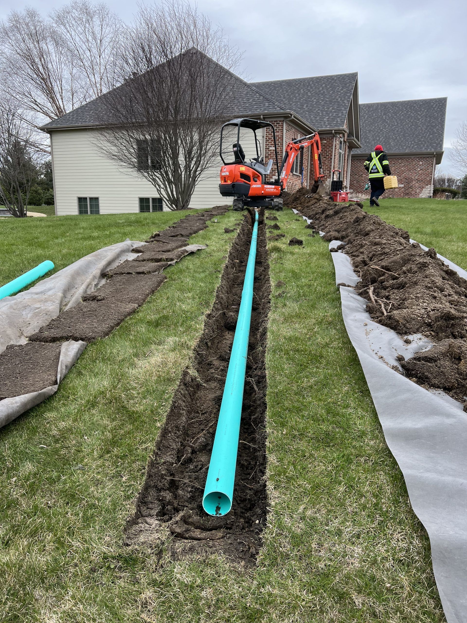 Construction site: Excavator in a lawn trench laying a turquoise pipe, near a house. Worker in safety vest.