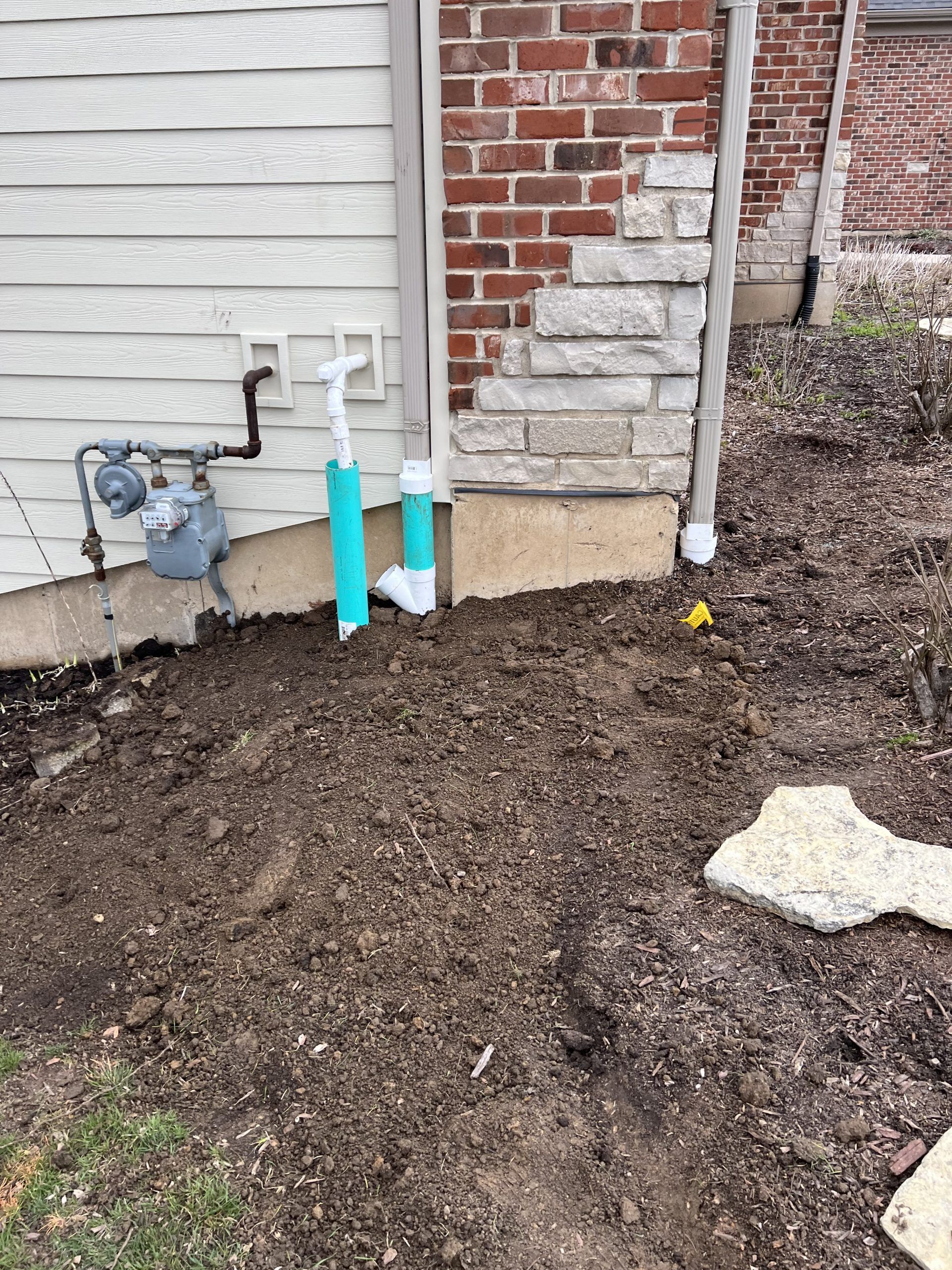 Dirt and partially buried pipes near a house with siding and brick, including a gas meter.