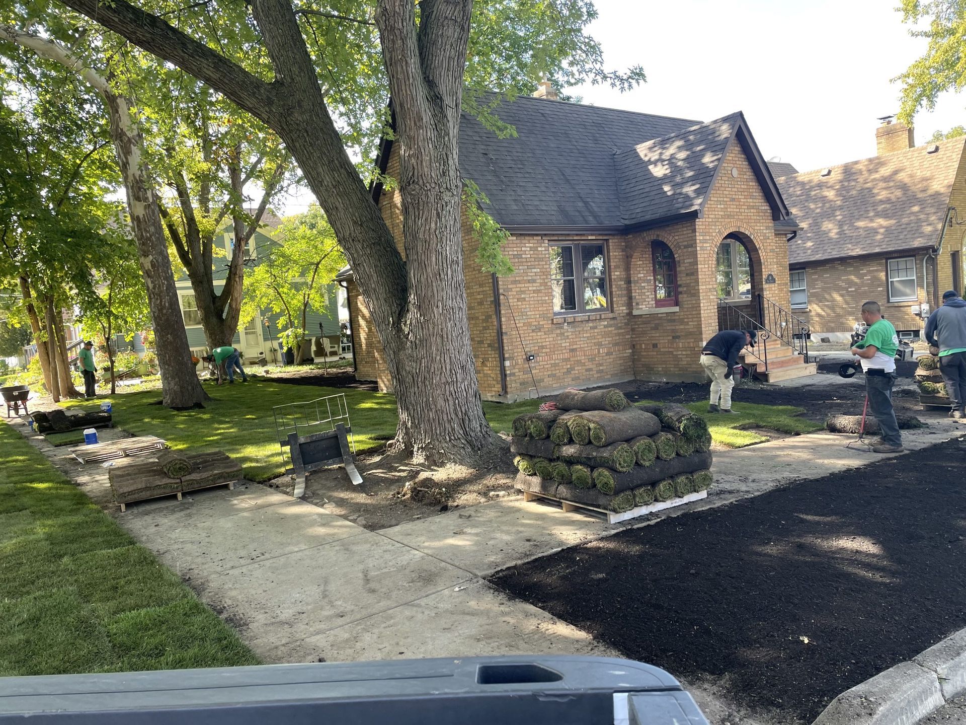 Workers installing sod in front yard of a brick house. Rolls of sod on a pallet, people working on the lawn.