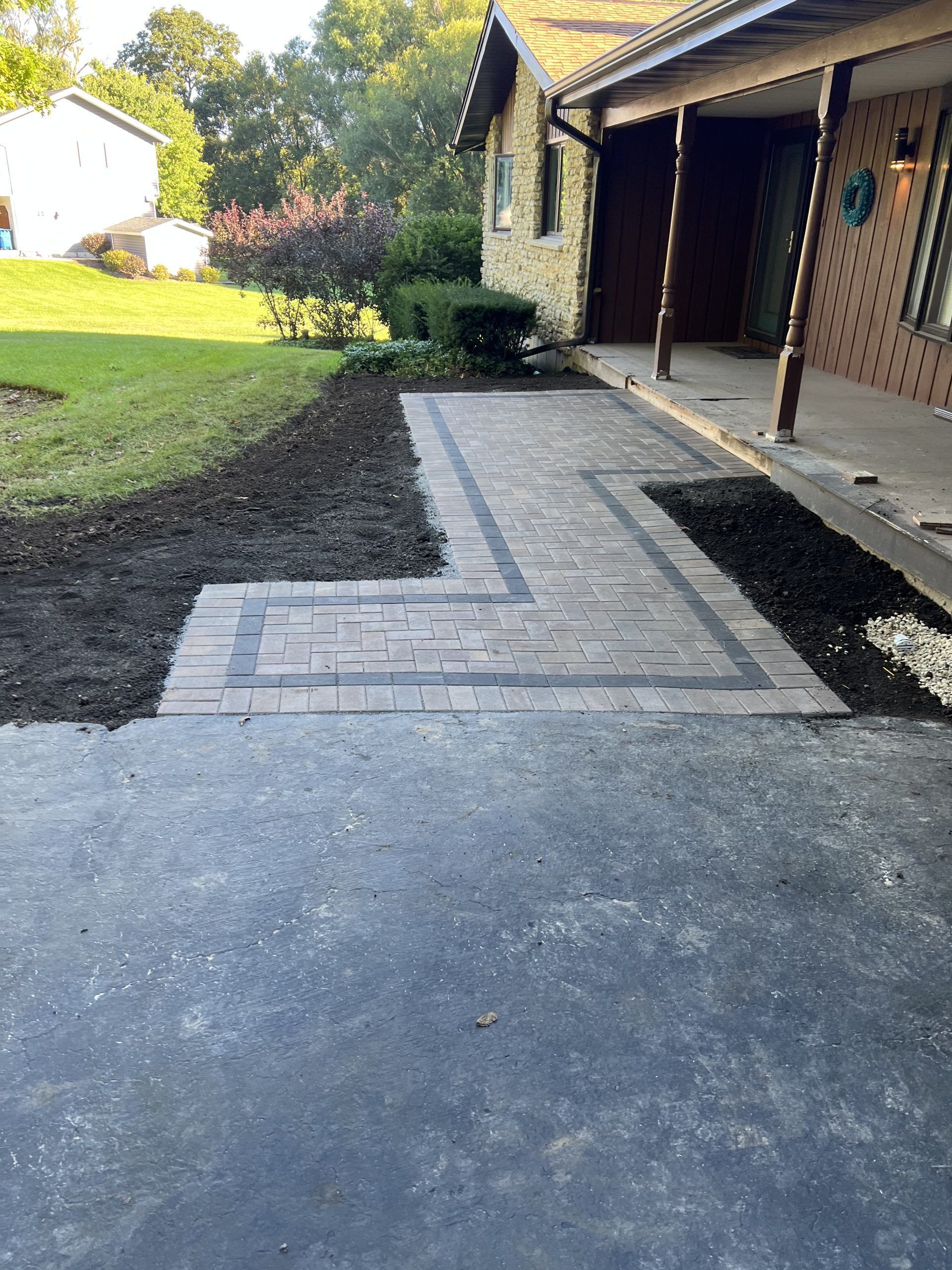 Brick pathway with dark border leads from driveway to a house with porch.