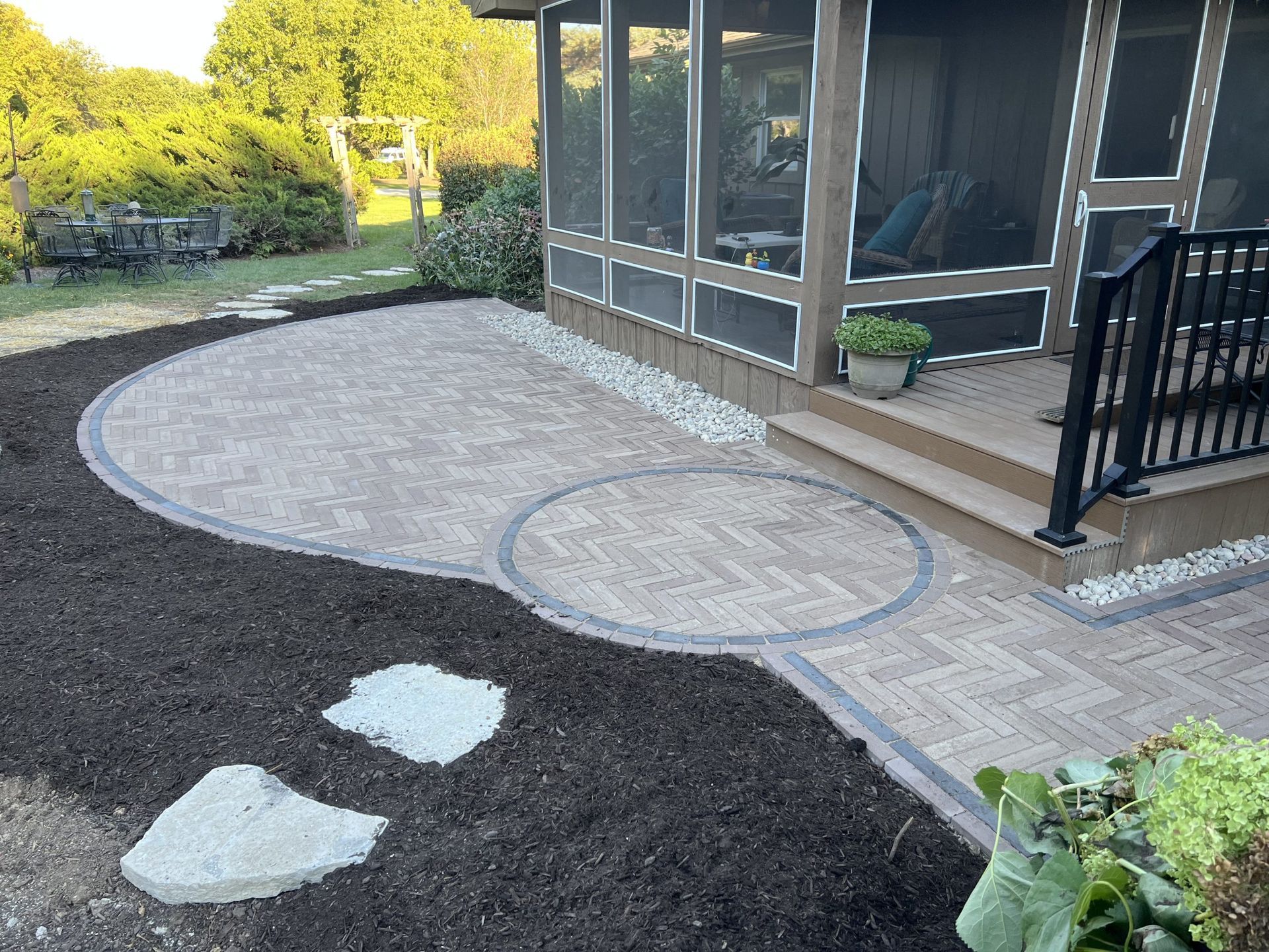 Paver patio with a circular design, leading to a screened porch entrance, surrounded by mulch and landscaping.
