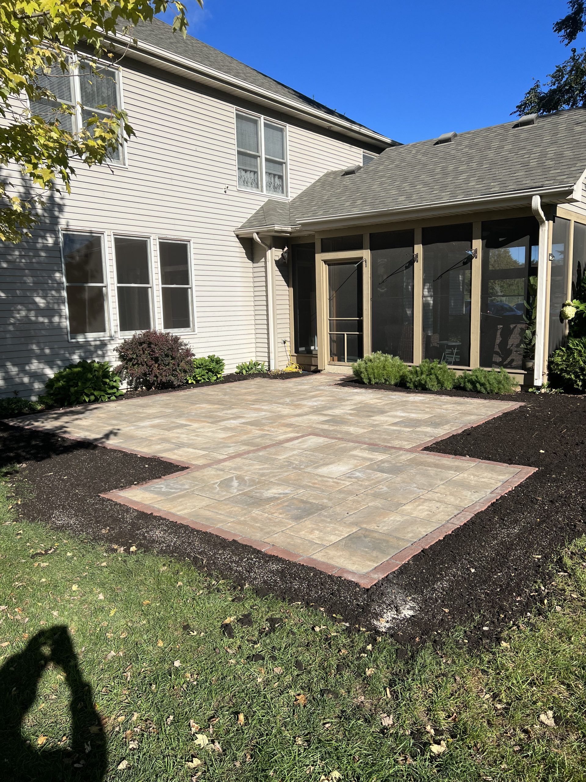 Backyard patio with pavers, surrounded by mulch and grass, next to a screened porch and house.