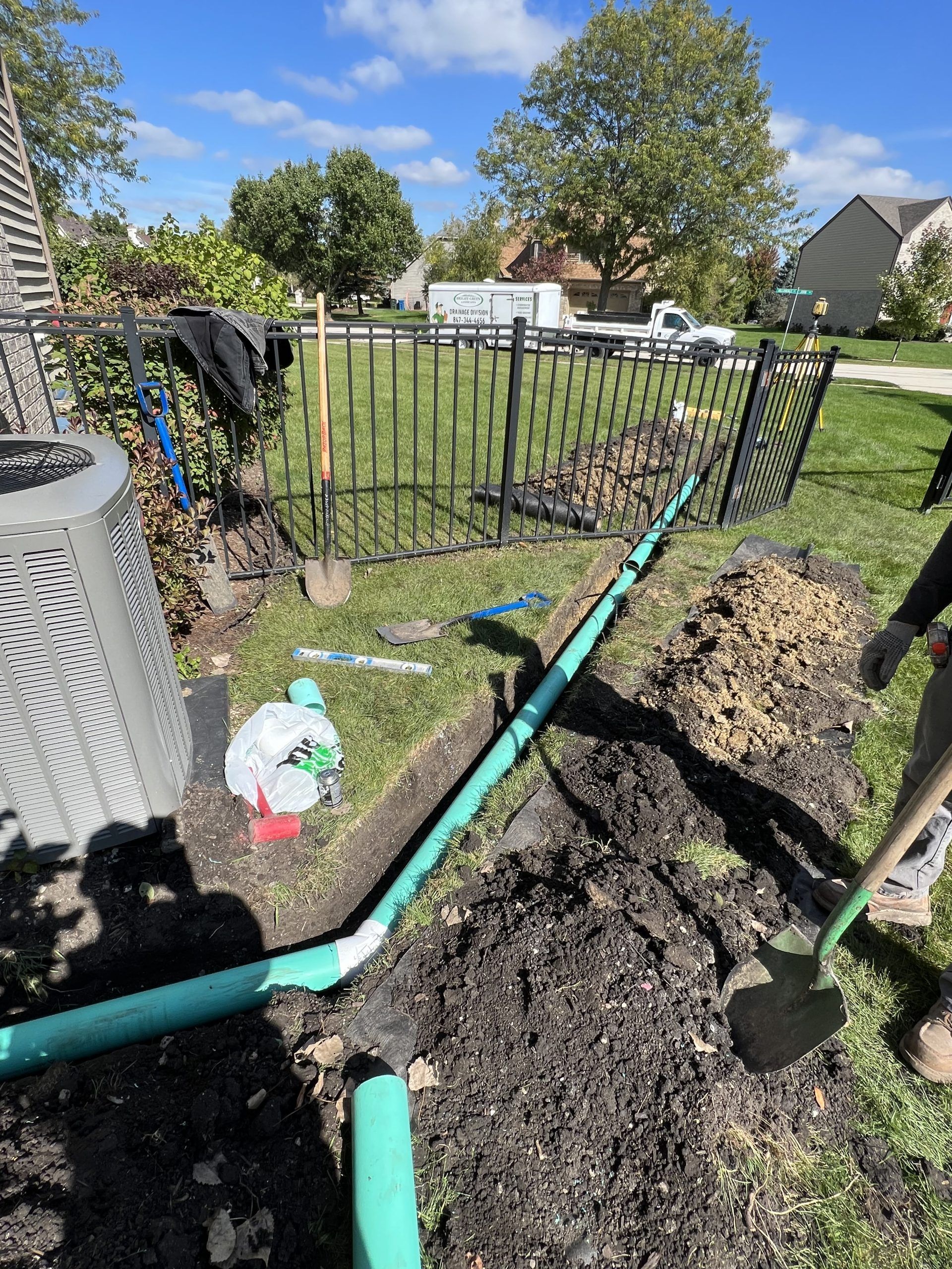 Installation of green drainage pipe in a yard, near a fence and dirt mounds, under a blue sky.