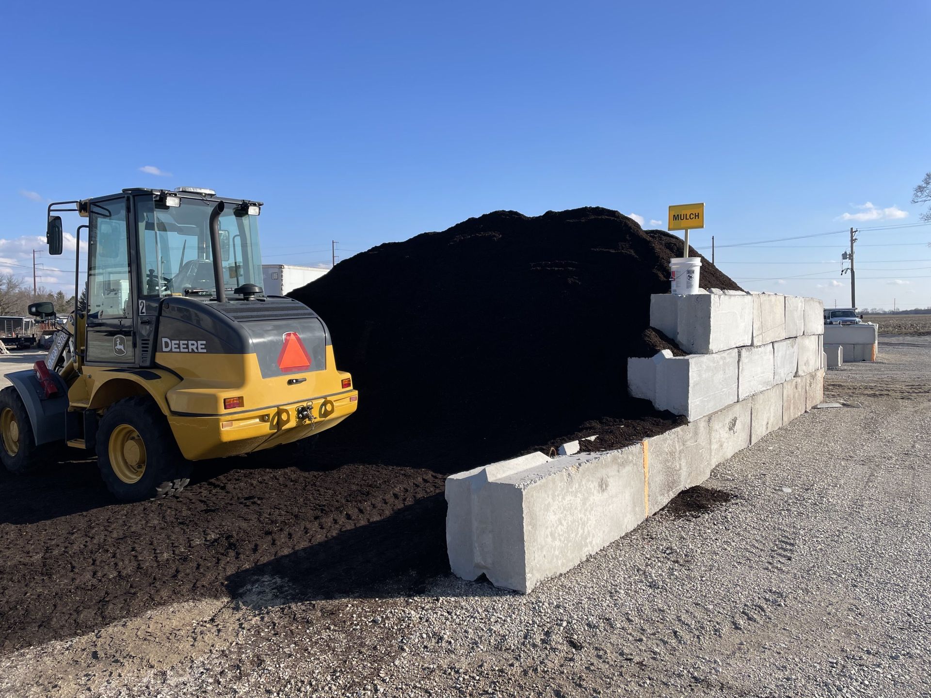 Yellow loader near a large pile of dark mulch next to concrete blocks, outdoors under a blue sky.