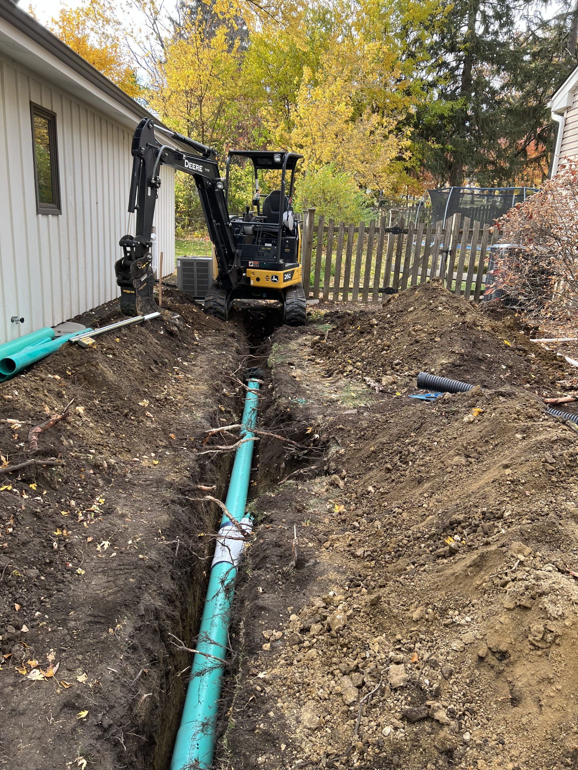 Mini excavator digging a trench, next to a house with blue drainage pipes laid in the trench.