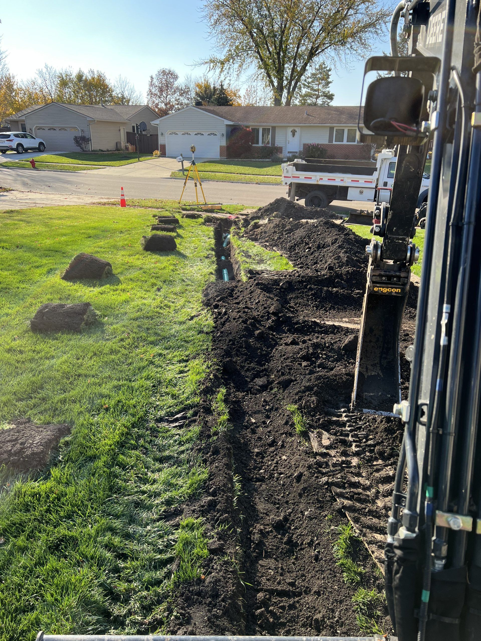 An excavator digging a trench in a grassy yard near a residential street.