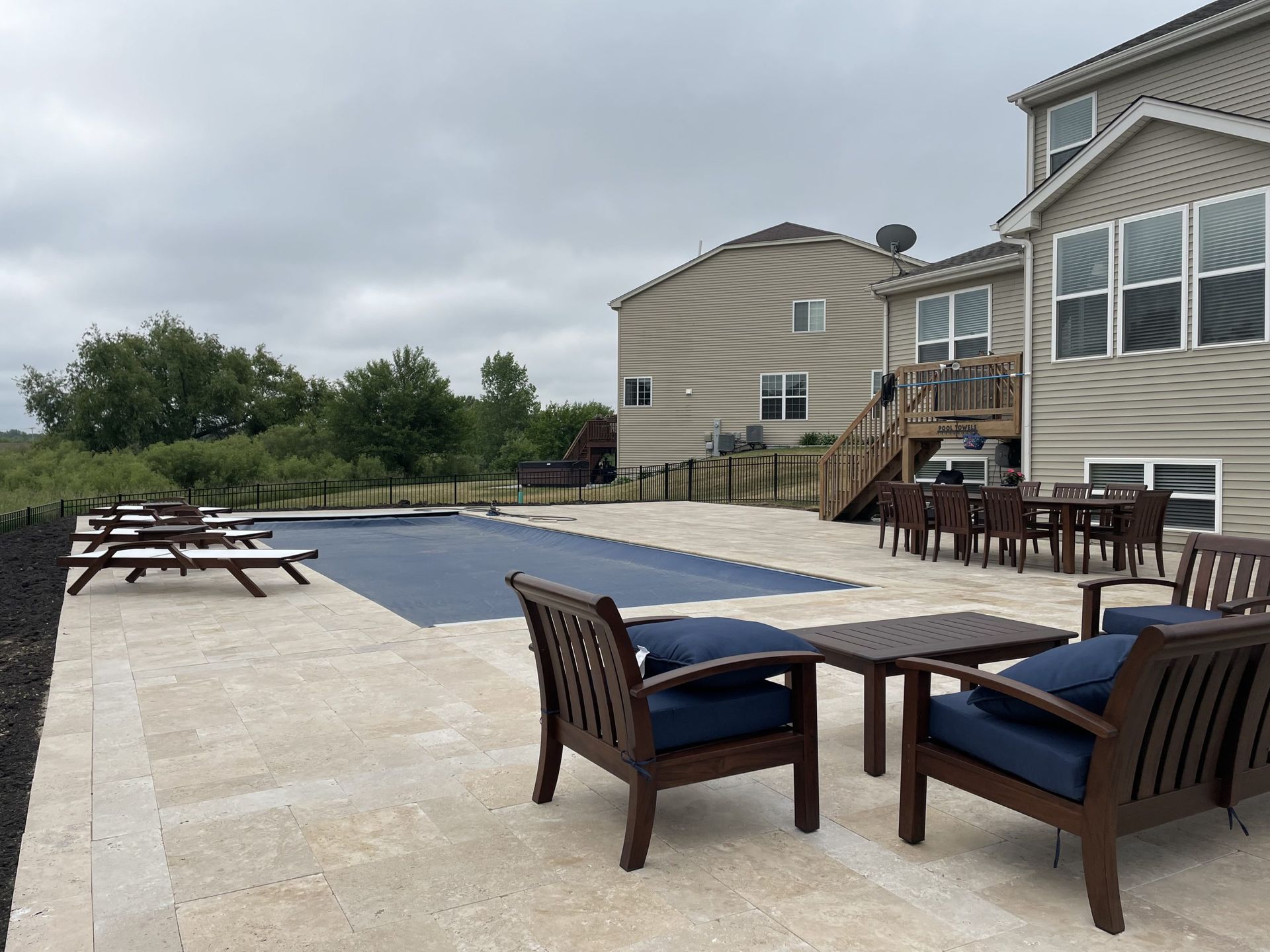 Patio with pool covered in blue; wooden chairs and table, house in background. Overcast day.
