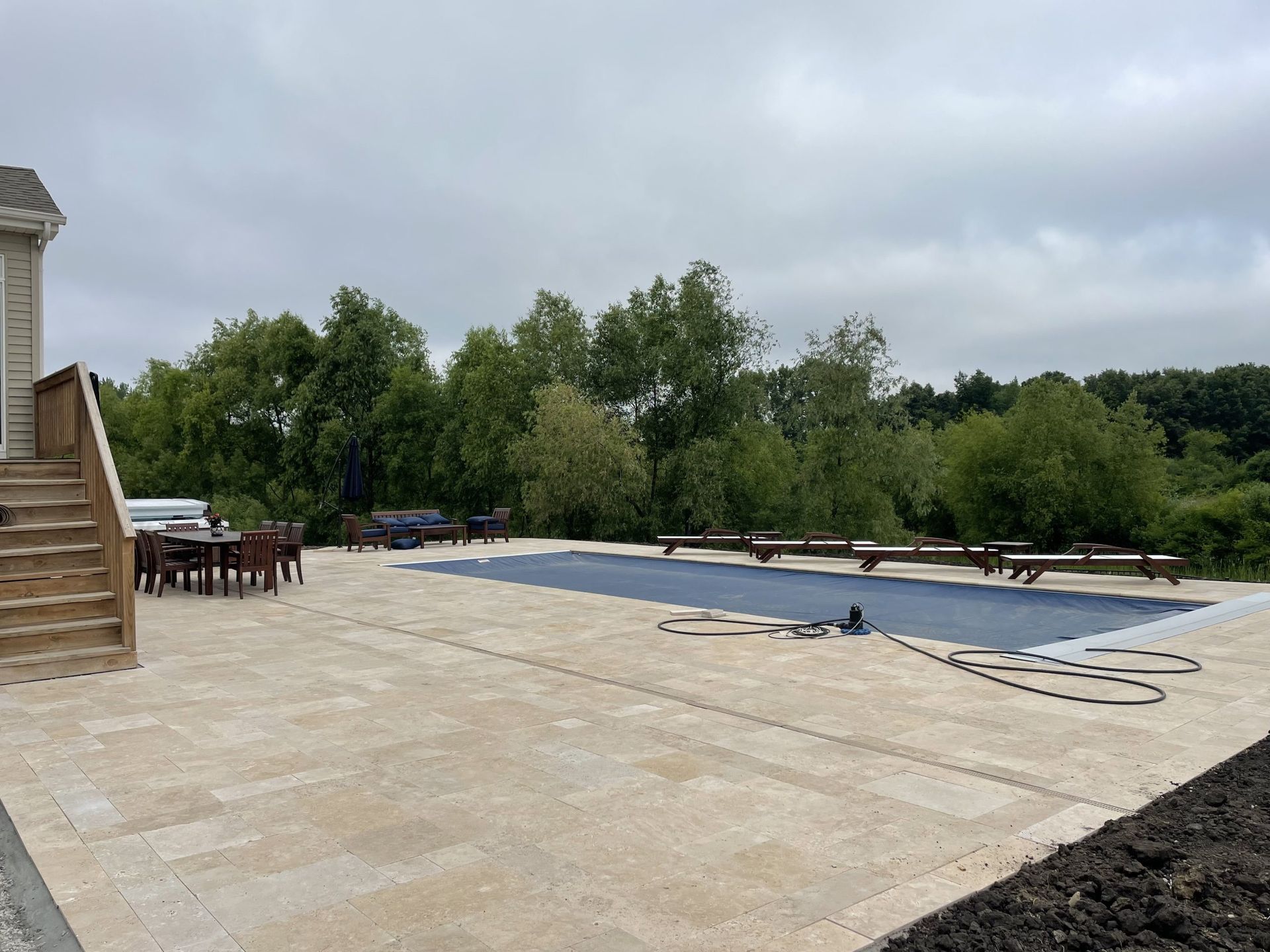 Patio with a pool covered by a blue tarp, wooden stairs on the left, trees in the background under an overcast sky.