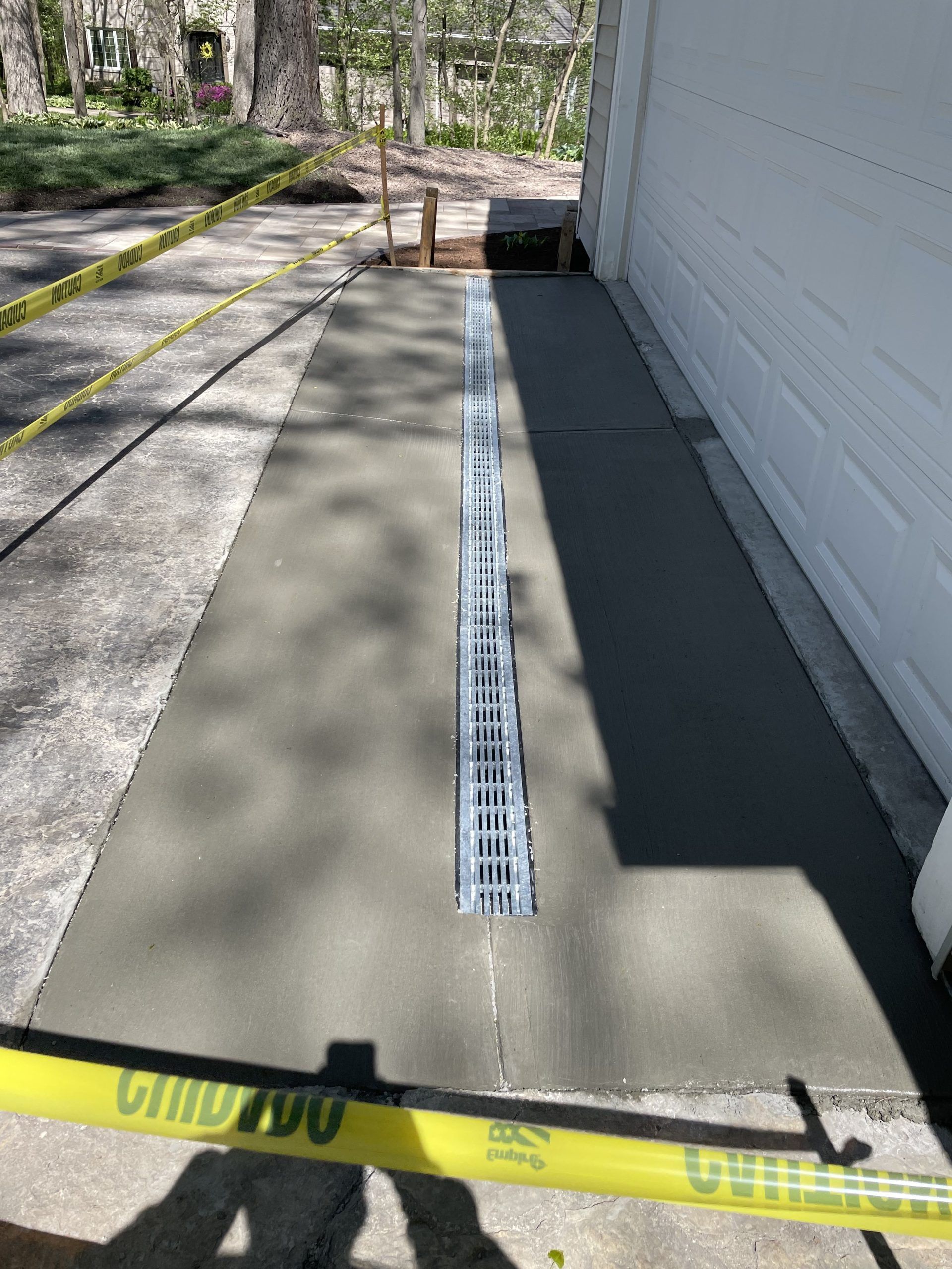 Newly poured concrete driveway with a central drain, next to a white garage door. Yellow caution tape.
