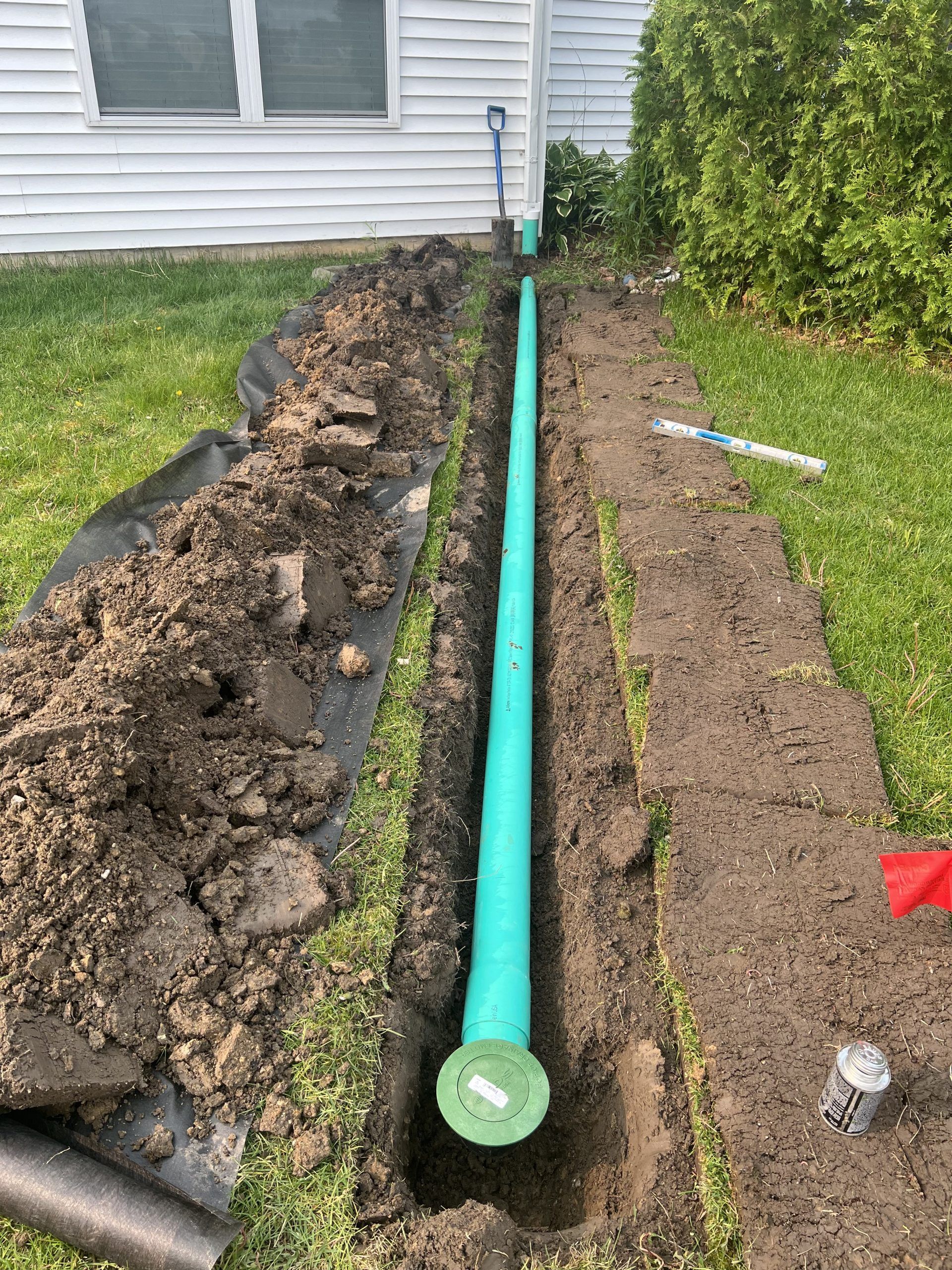 Trench with green pipe installed along a house foundation; dirt, shovel, and building in the background.