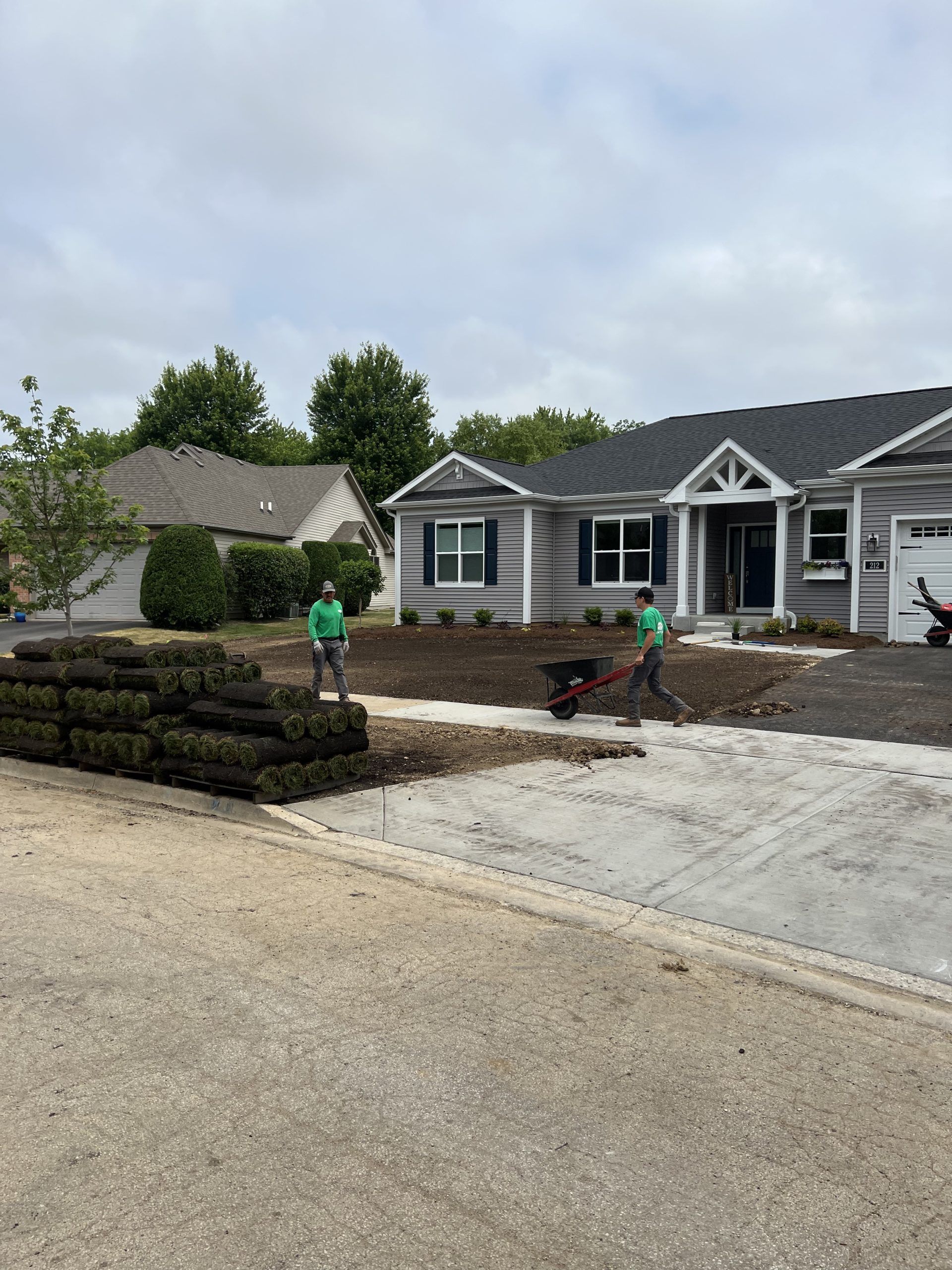 Two people laying sod in front of a gray house. Sod stacks on the left. Cloudy sky.