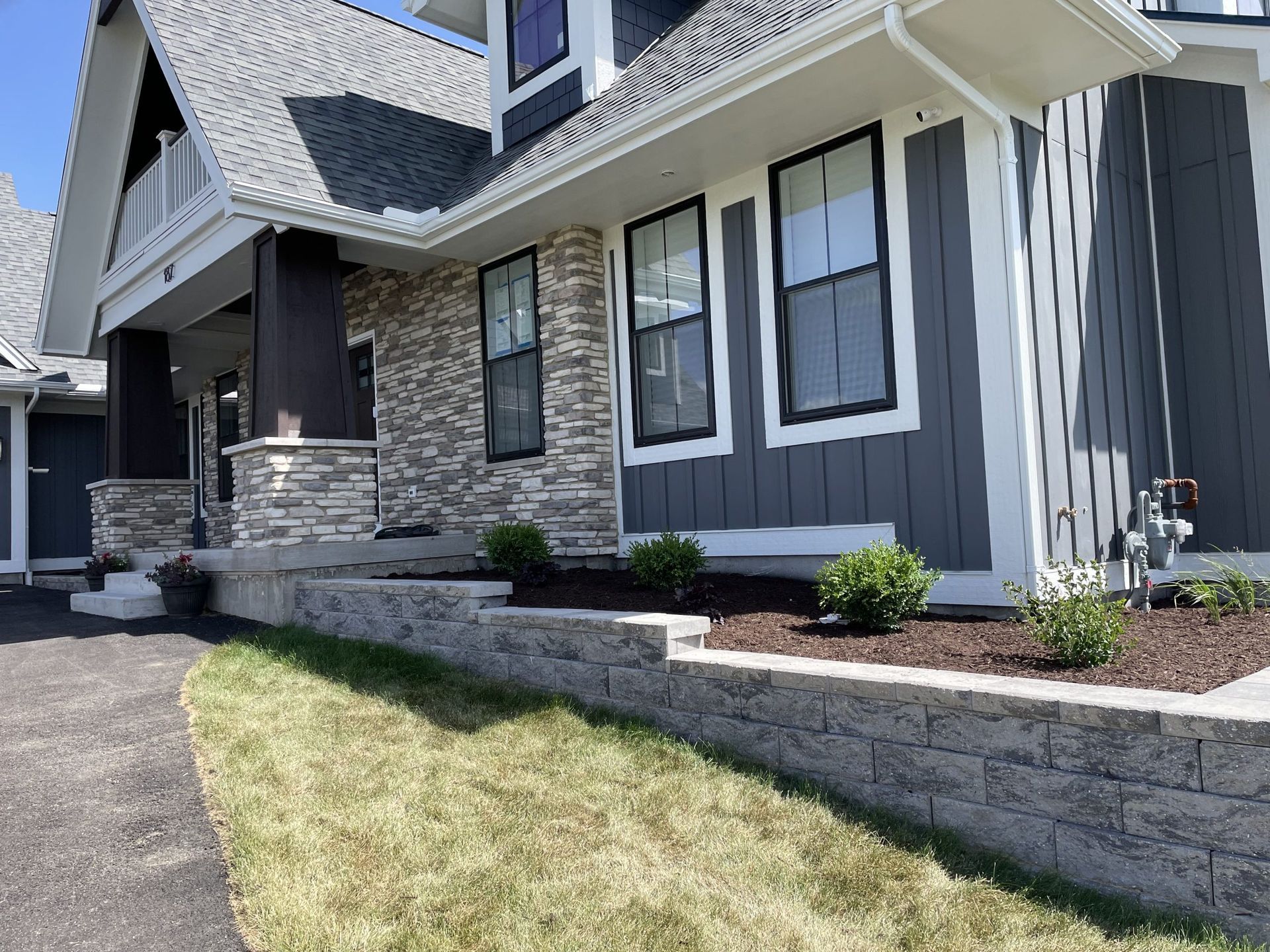 Gray house with stone and siding, a retaining wall, and green lawn on a sunny day.