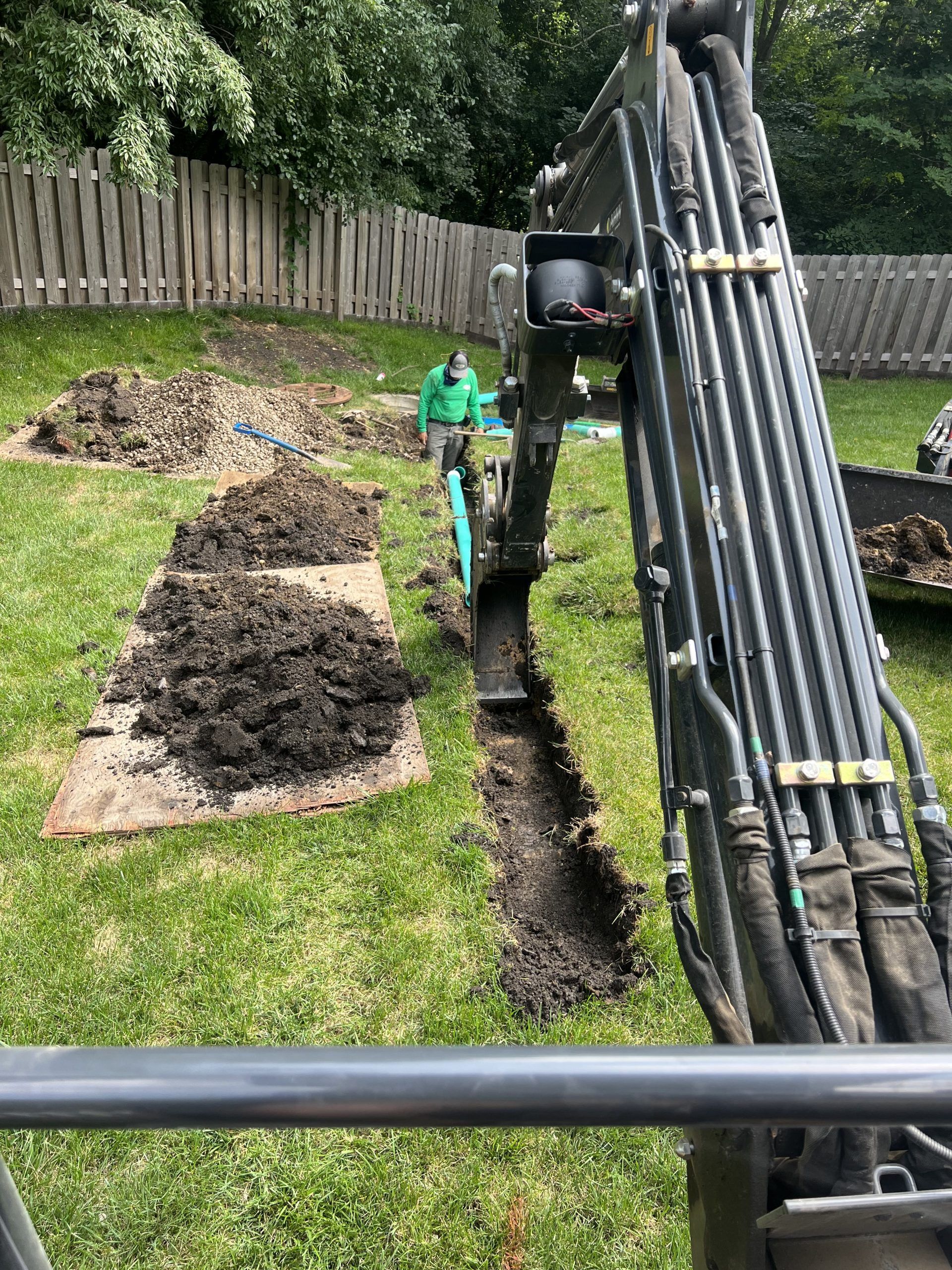 An excavator digging a trench in a grassy backyard. A person stands nearby. Brown soil and a wooden fence are visible.