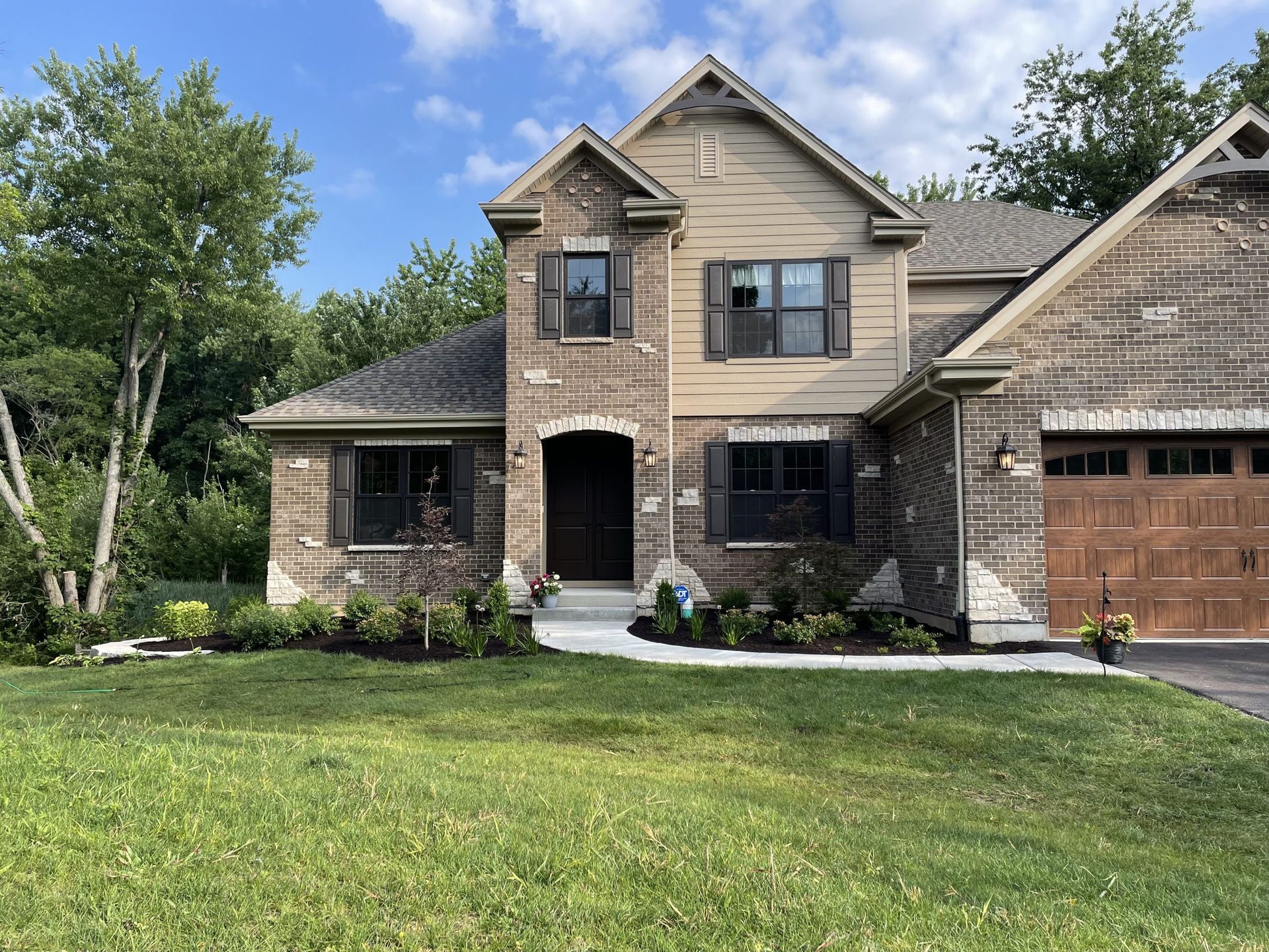 Two-story brick house with brown garage door, black shutters, and well-manicured lawn under a blue sky.