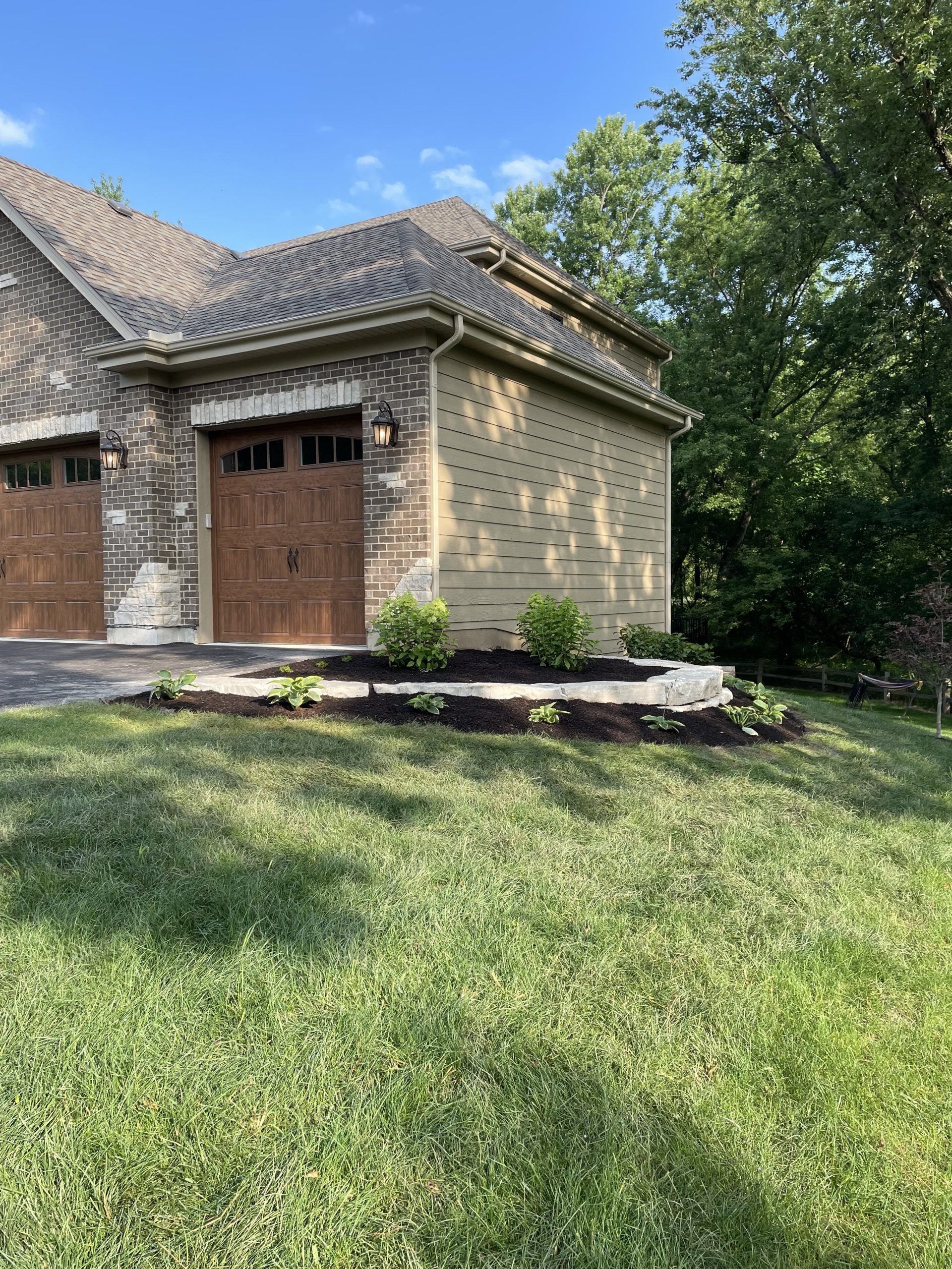 Brick garage with brown doors, landscaping, and green lawn on a sunny day.