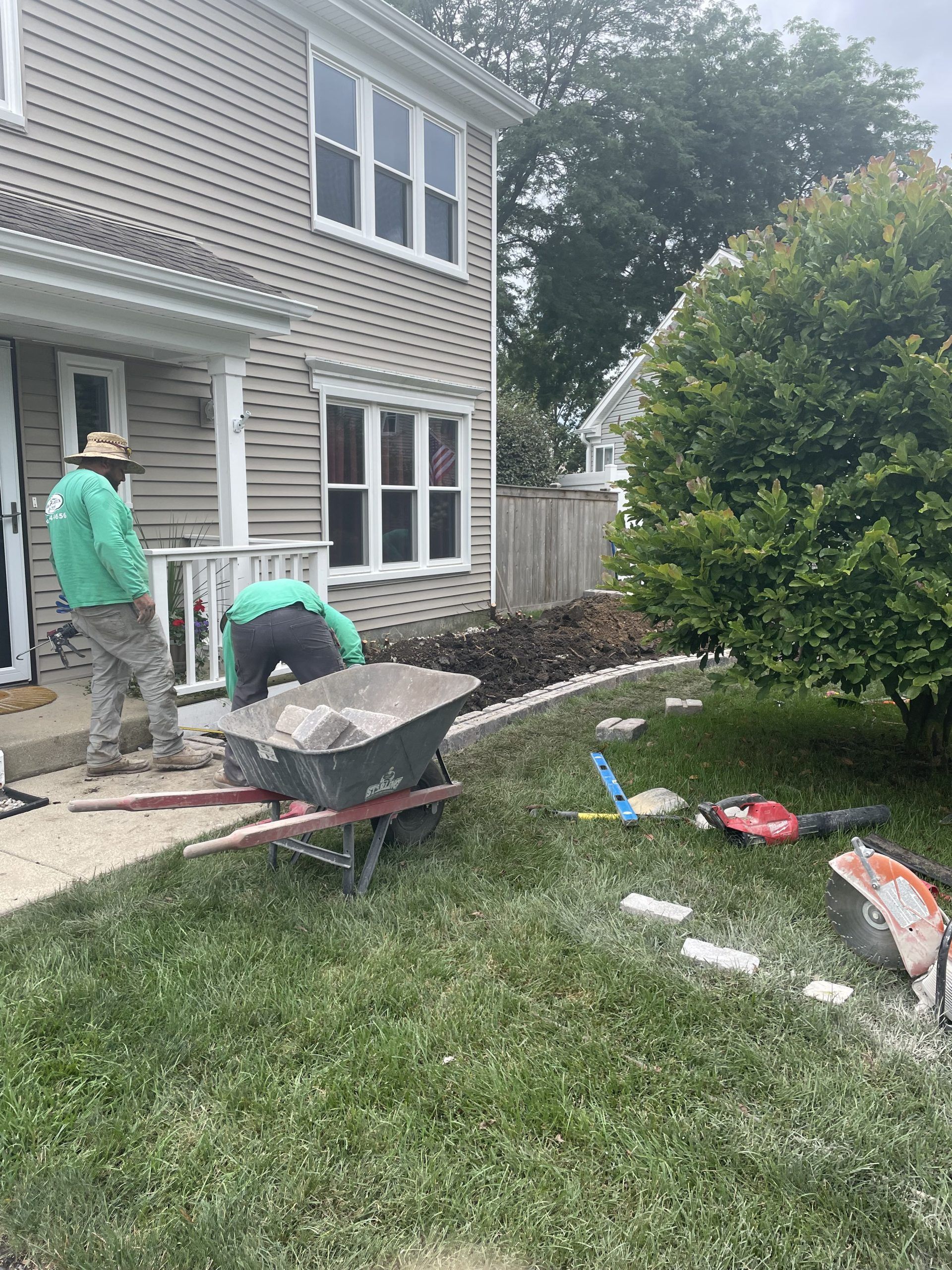 Workers landscaping near a house, wheelbarrow with rocks, stepping stones, grass, trees.