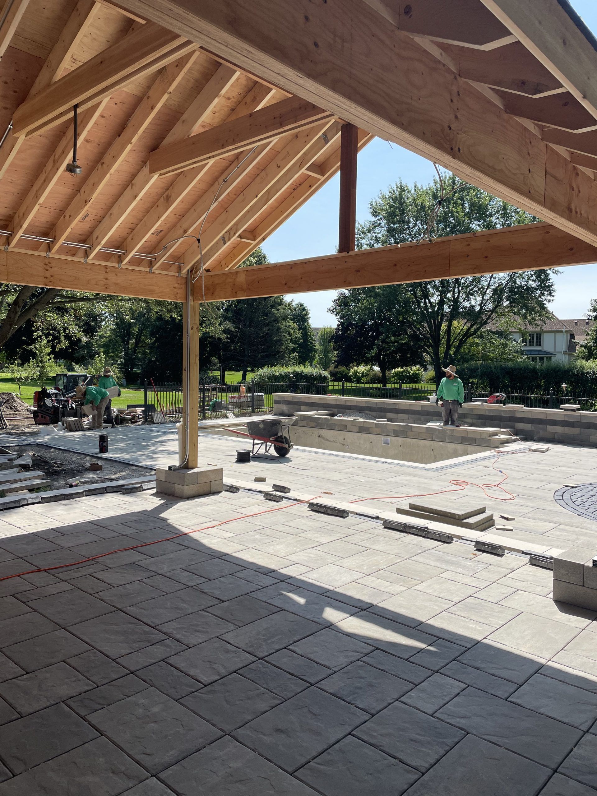 Wooden pavilion under construction; pavers, workers, and a pool in a sunny outdoor setting.