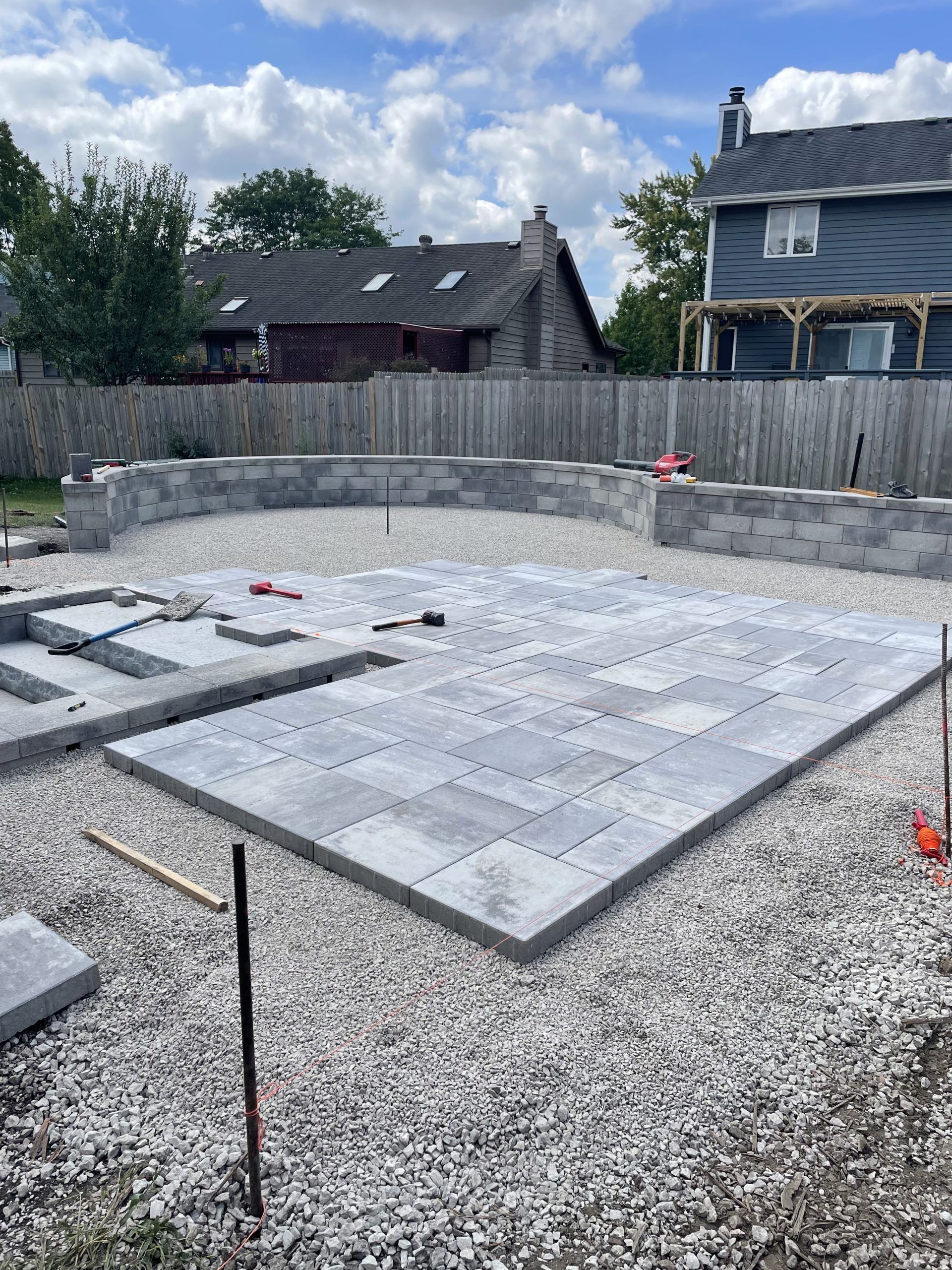 Construction site with gray pavers being laid for a patio. Gray retaining wall in the background. Gravel and sky.