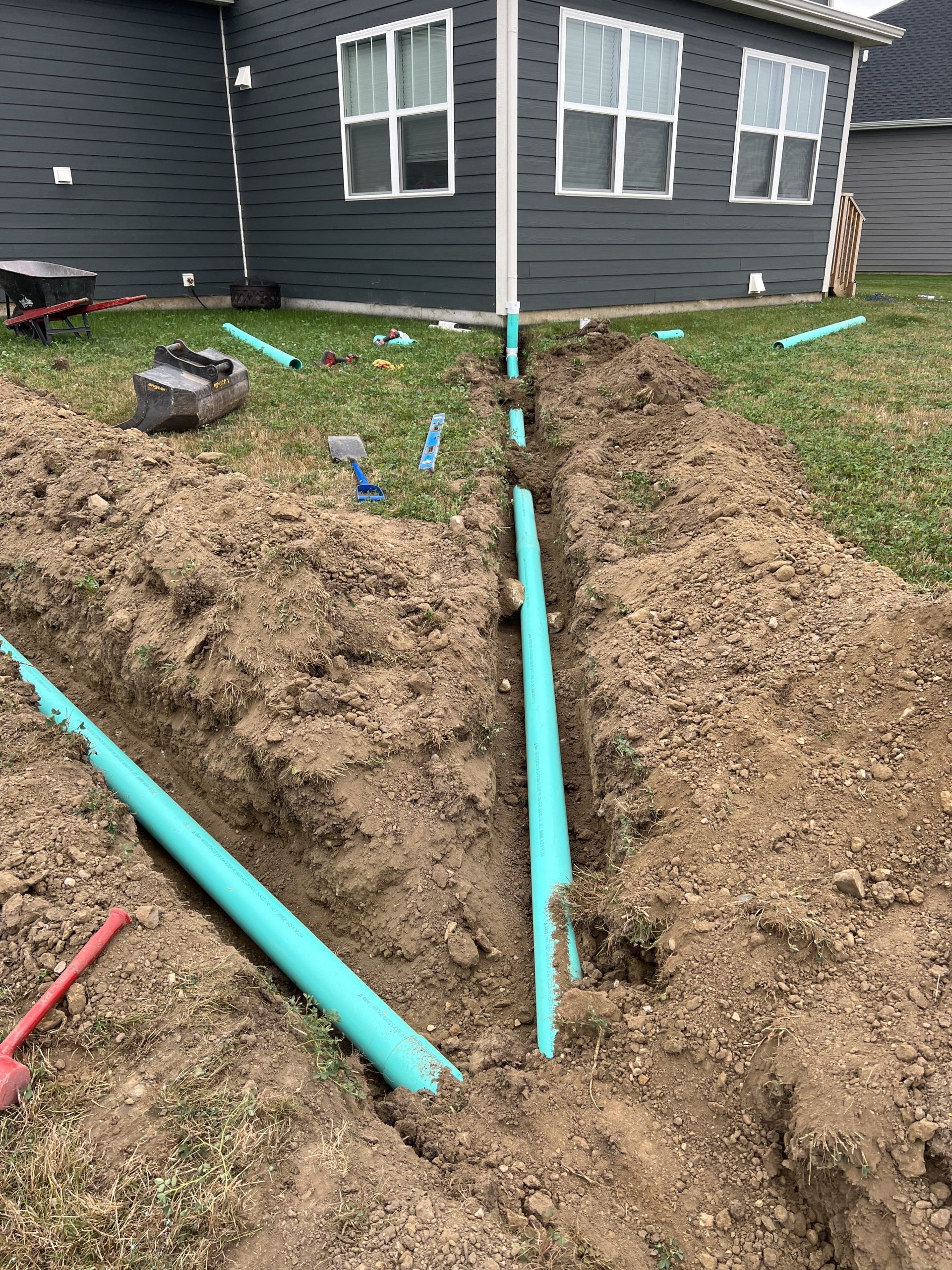 Trenches in yard with blue PVC pipes laid, near a gray house with white windows.
