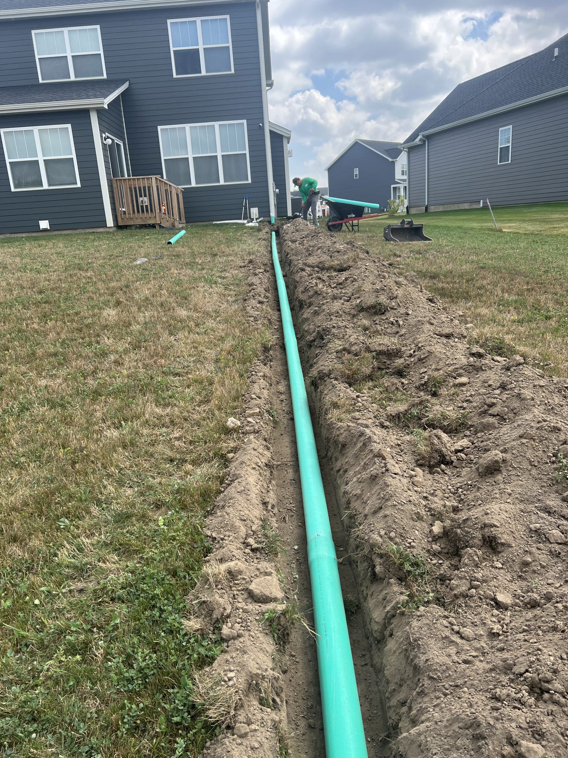 A green pipe in a trench runs through a grassy yard near houses.