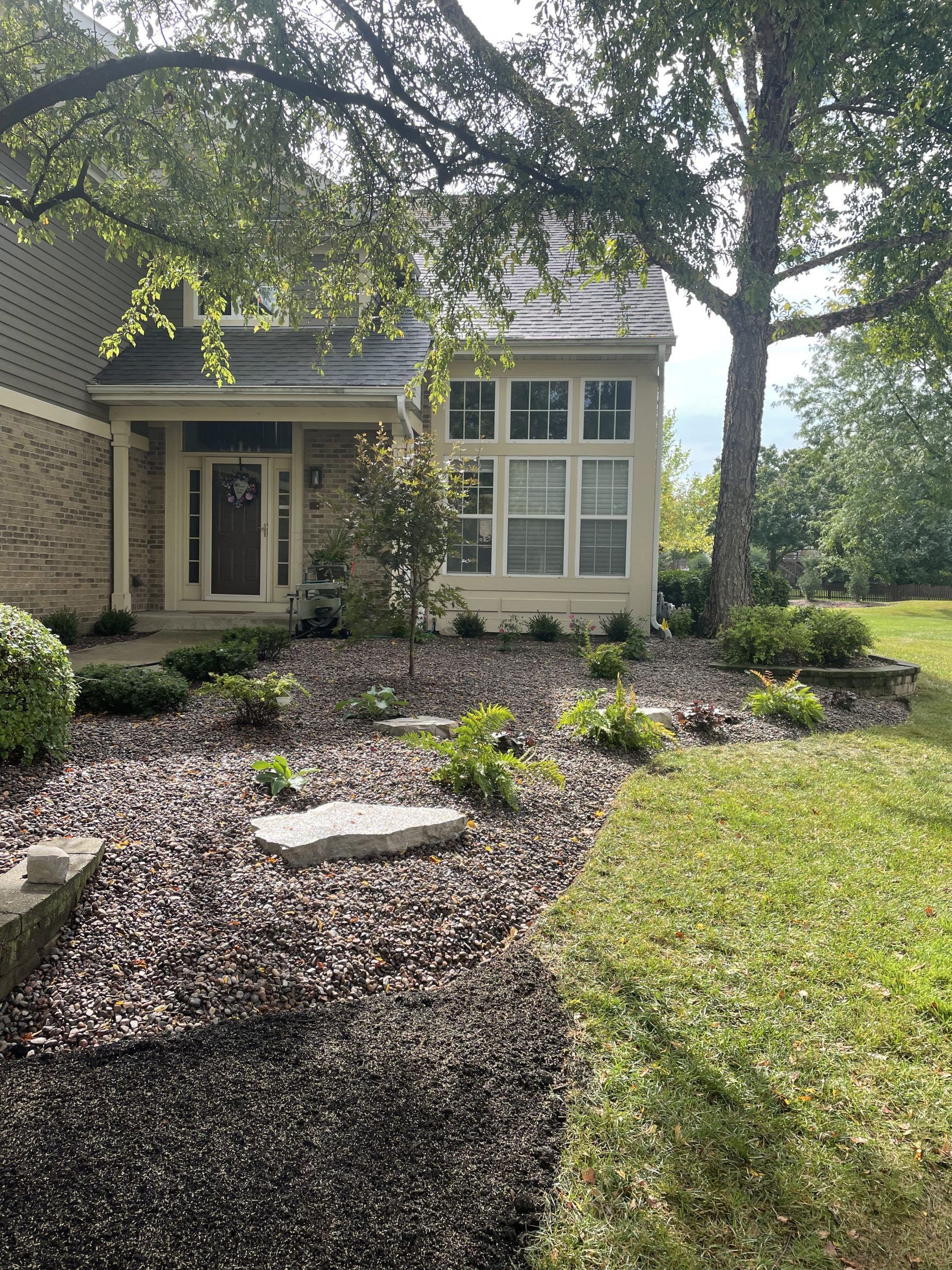 Beige house with porch, windows, and landscaping. Dark mulch with stepping stones, bordered by green lawn.