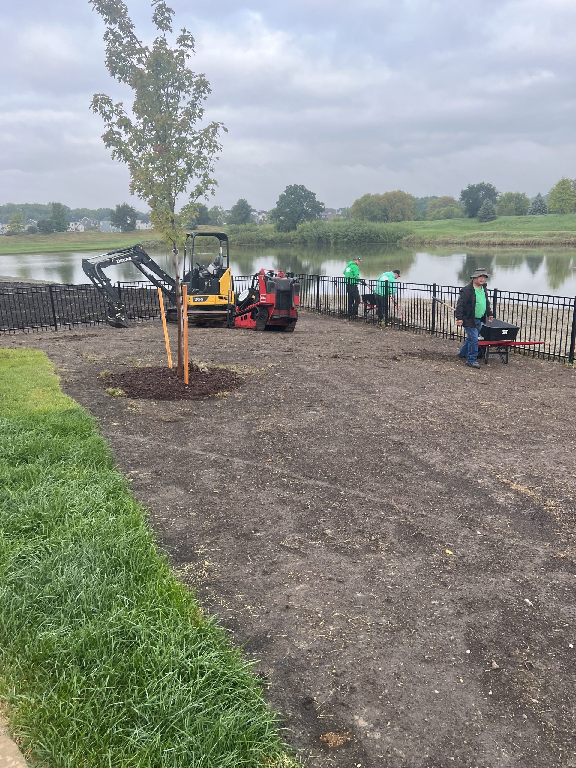 Workers planting saplings near a lake. Soil, small machinery, and bundled plants are present. Overcast sky.