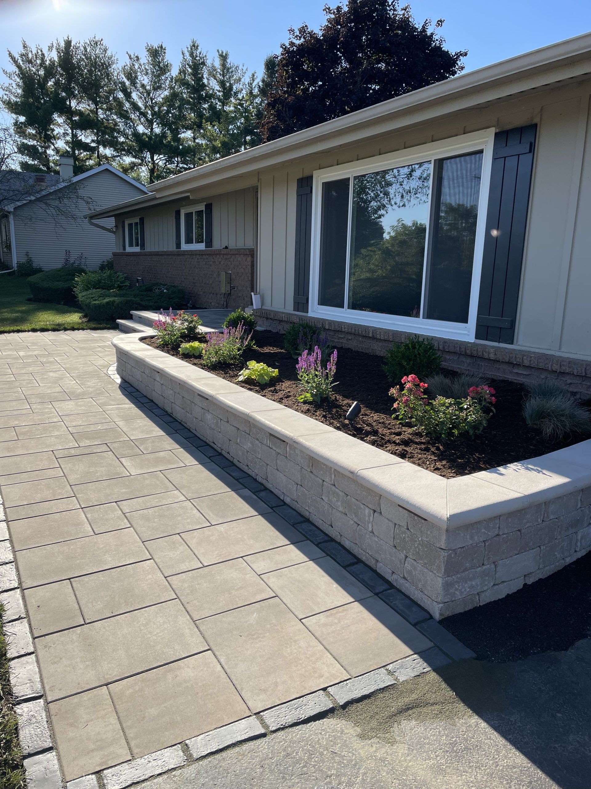 Exterior view of a beige house with a stone pathway and a raised flower bed filled with plants.