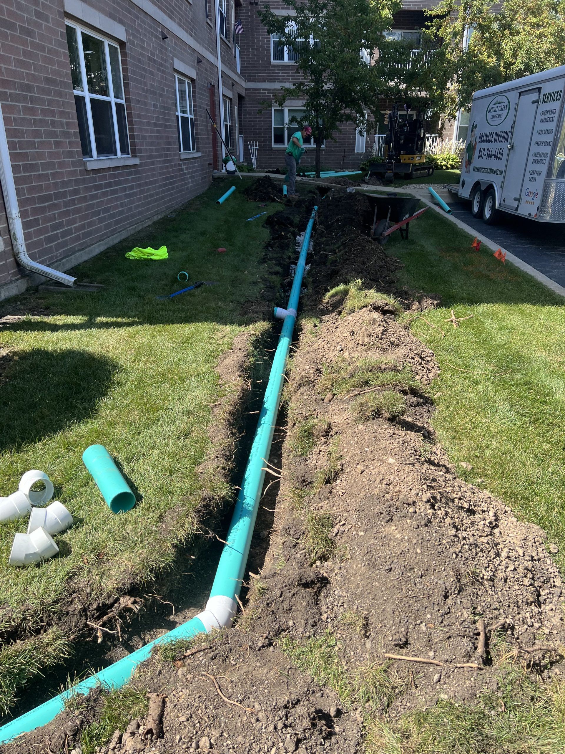 A trench with green pipes being installed in a grassy area next to a brick building.