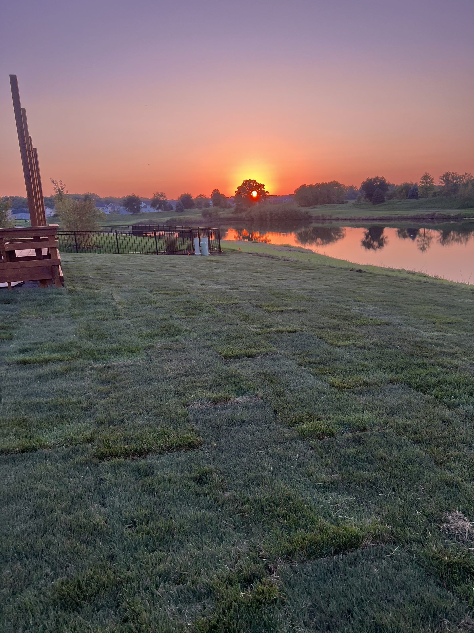 Sunrise over a calm lake, with green grass in the foreground and a wooden structure on the left.