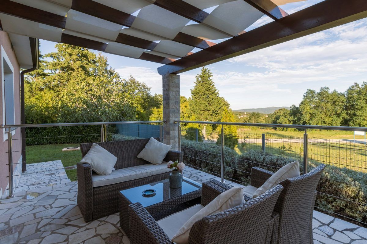 Patio with wicker furniture, shaded by a canopy, overlooking a green landscape.