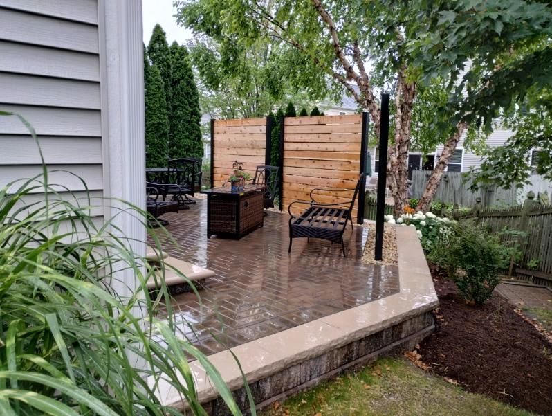 Patio with brick pavers, wooden privacy fence, black metal furniture, and landscaping.