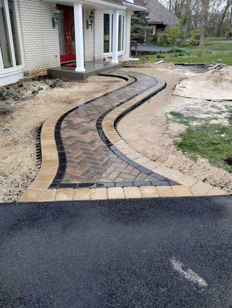Brick pathway leading to a house entrance. Brown and black bricks, curving design, set in sand.