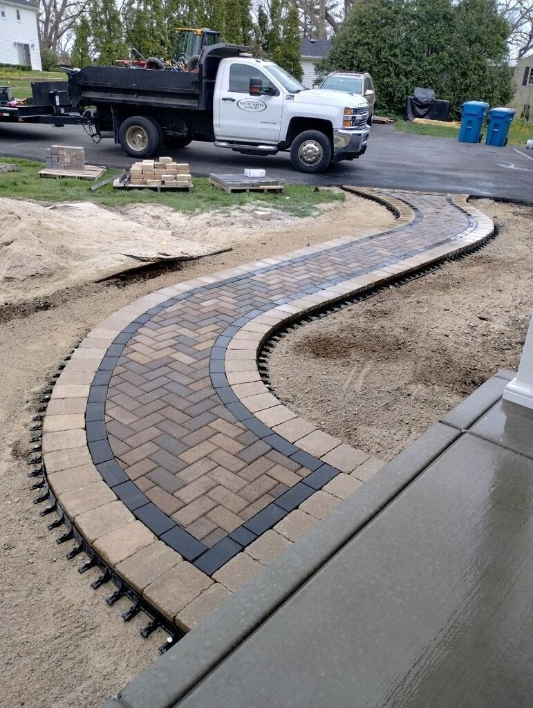 Curving brick pathway with a dark border, under construction, with a dump truck in the background.