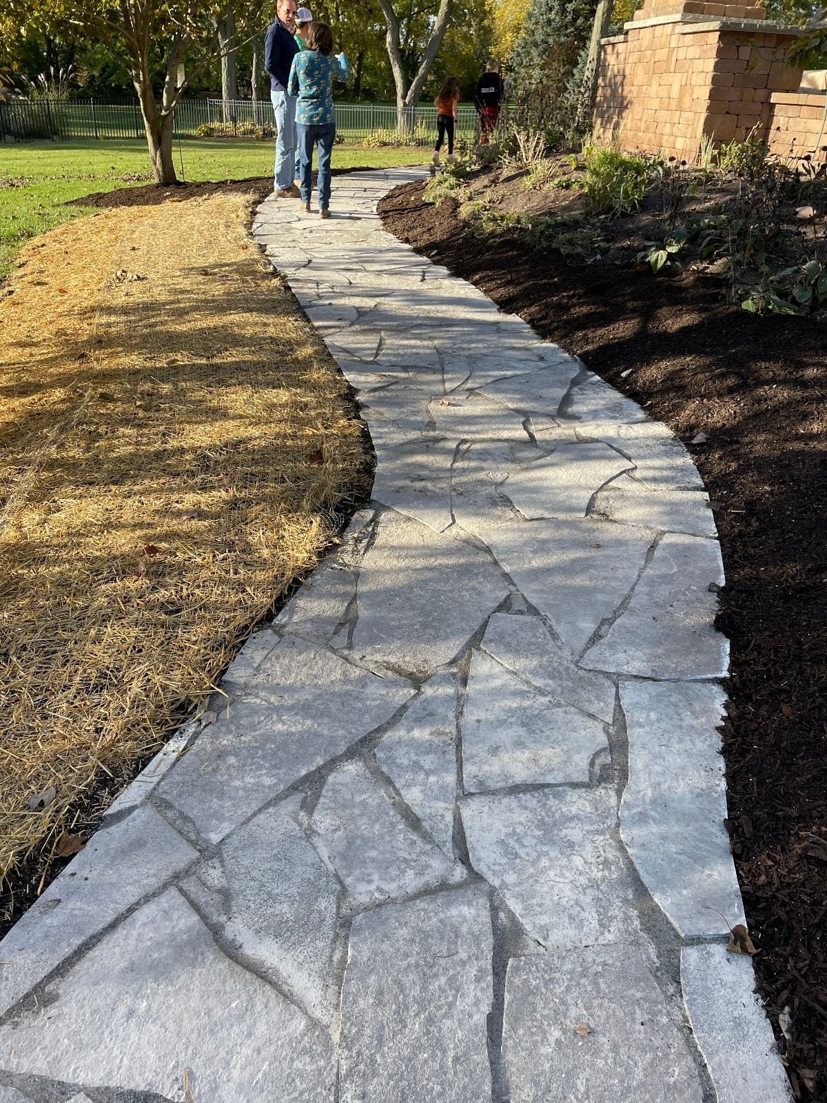 Stone path winds through a yard, two people walk towards the end. Brown mulch and grass border the path.