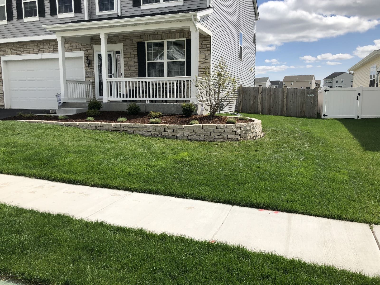 Suburban house with a stone retaining wall and well-manicured lawn next to a sidewalk on a sunny day.