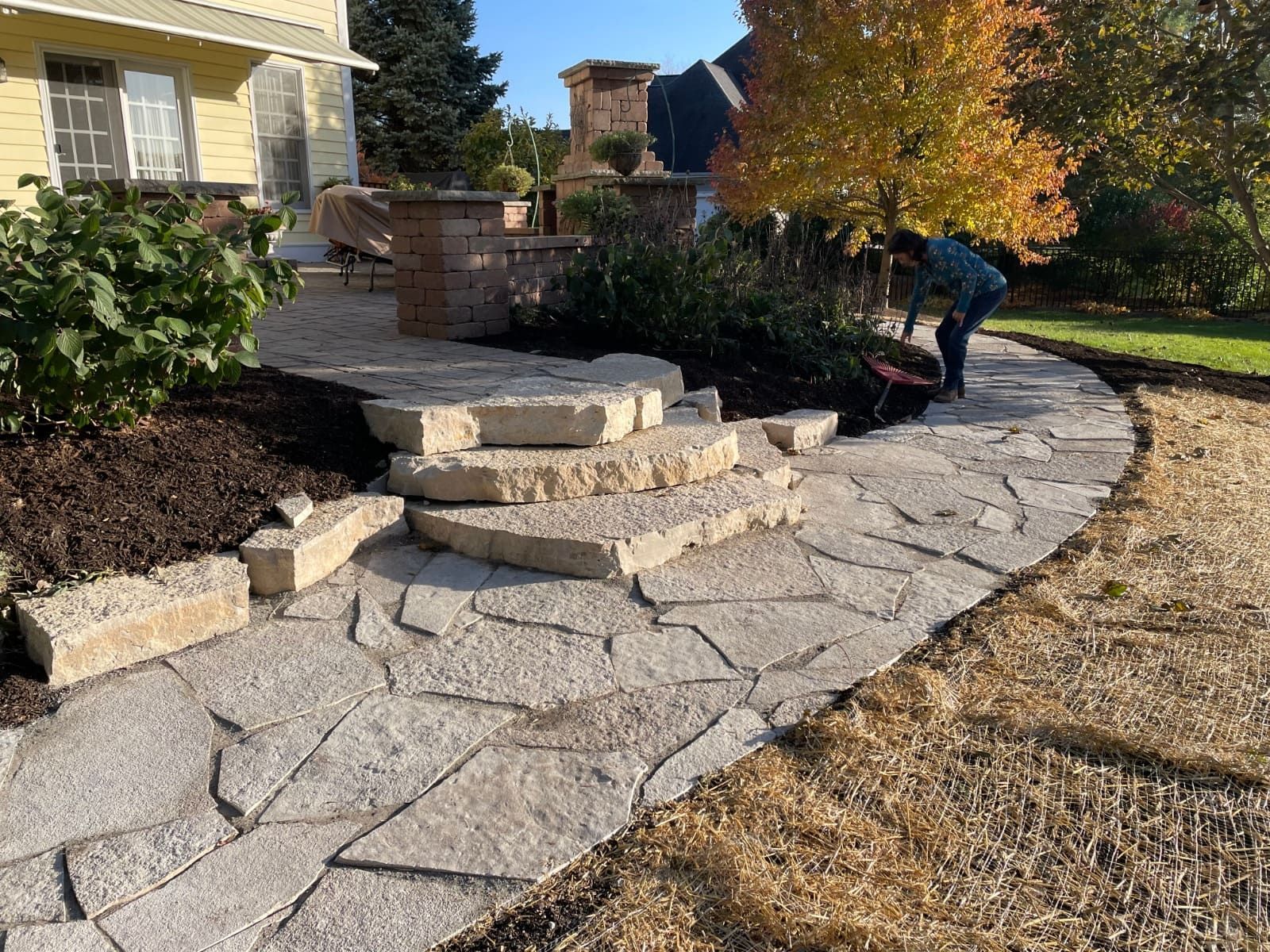Stone pathway with steps leading to a landscaped area; person tending to plants on the side; fall foliage.