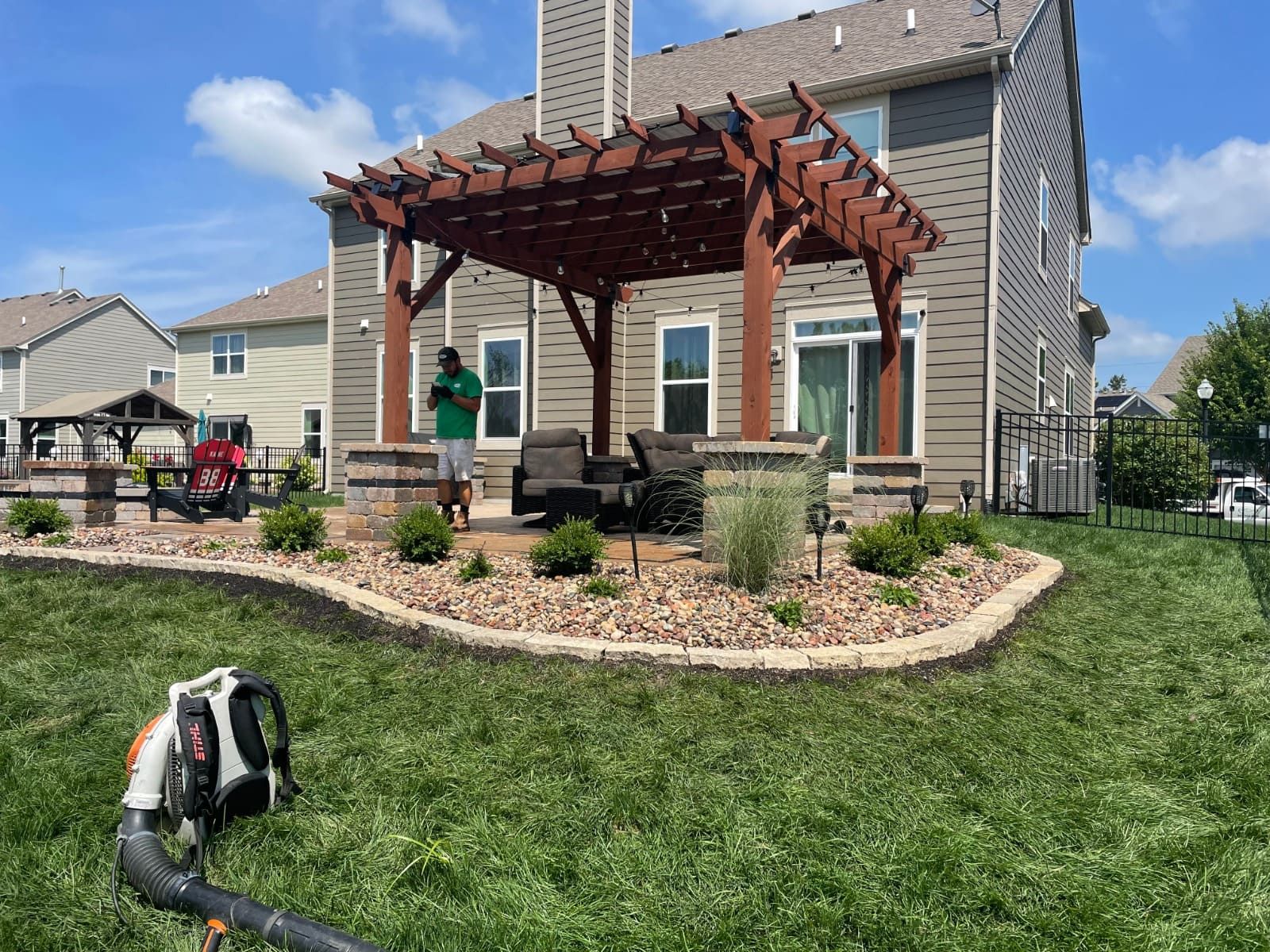 A backyard patio with a pergola, landscaping, and a person using a leaf blower on the grass.