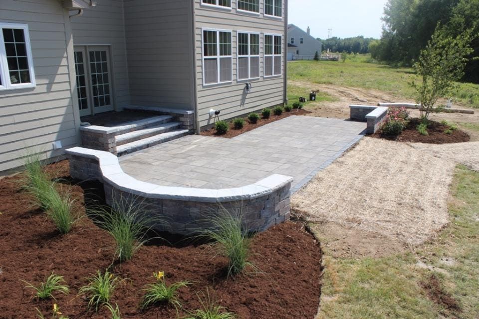 A brick patio with retaining walls extends from a house with steps. Mulched flower beds and tall grasses are in front.
