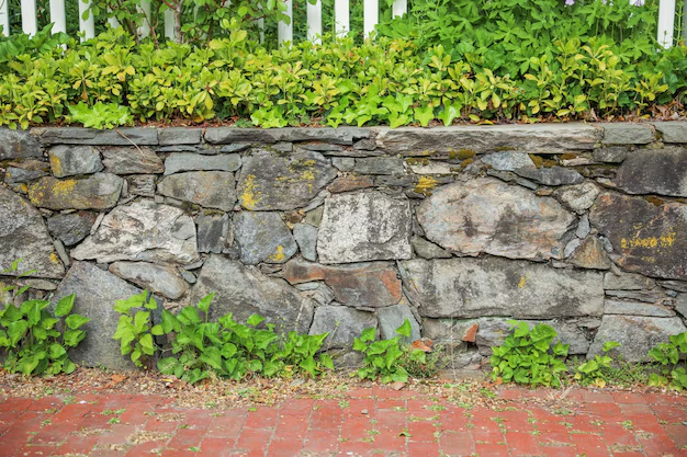 Stone wall with green plants, red brick below, and white fence with foliage above.