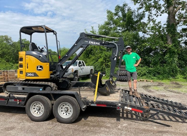 Man standing on a trailer with a John Deere excavator. Green shirt, trees in the background.