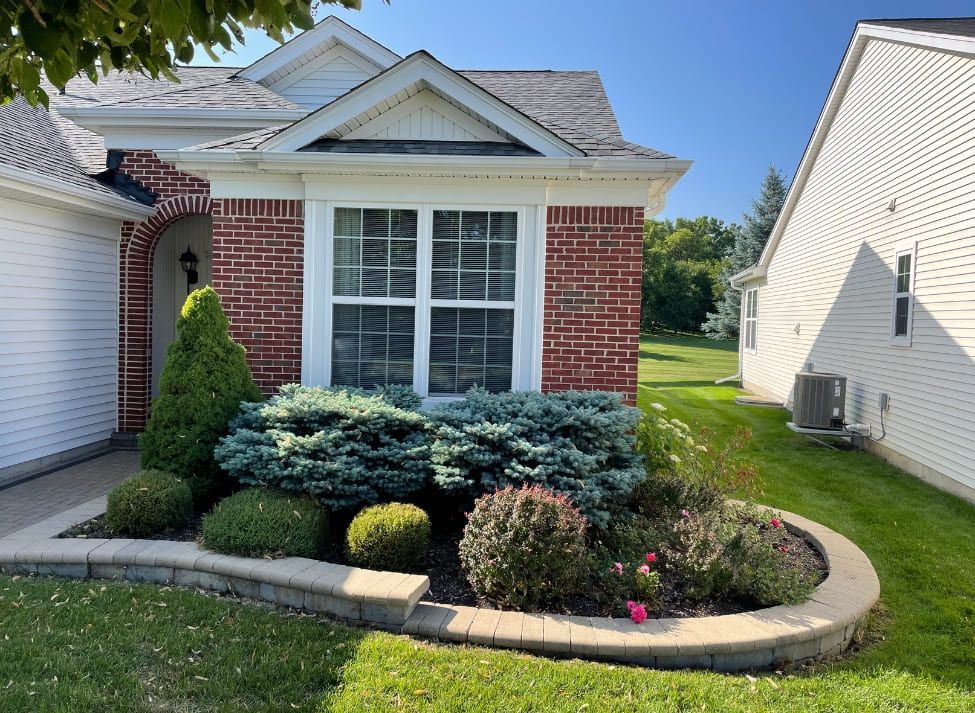 Brick house with flower bed of shrubs and bushes. Window and garage visible. Green grass, blue sky.