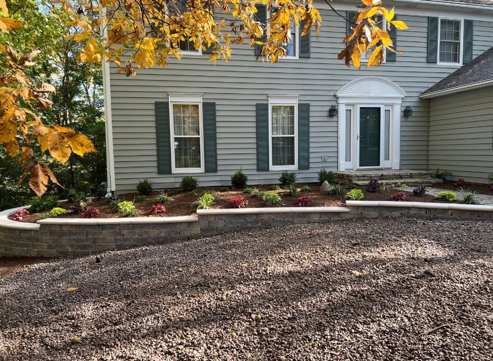 Two-story house with gray siding, a stone retaining wall, and a gravel driveway. Landscaped front yard with colorful plants.