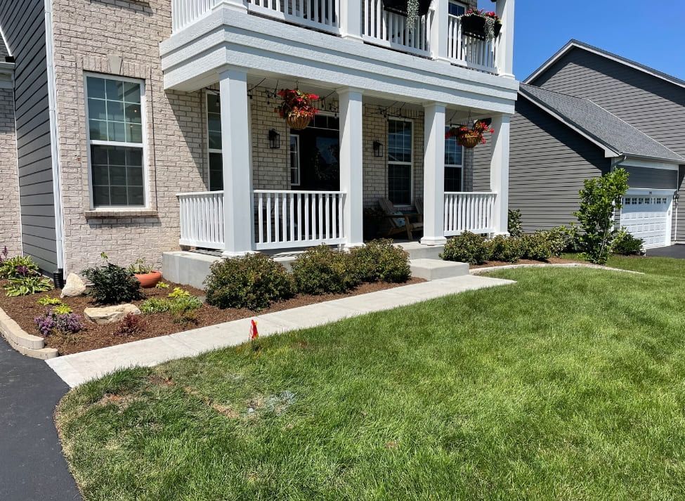 A brick house with a porch. White pillars and railing, lush green lawn, and blue sky.