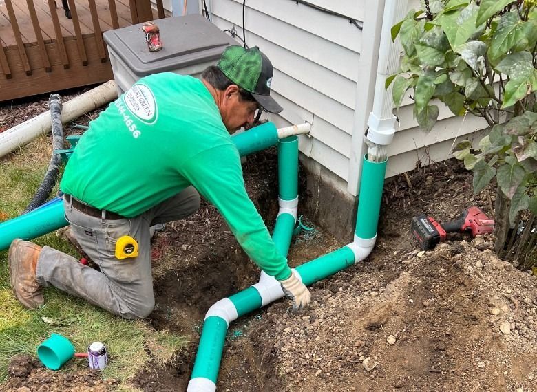 Man in green shirt installing drainage pipes near a house.