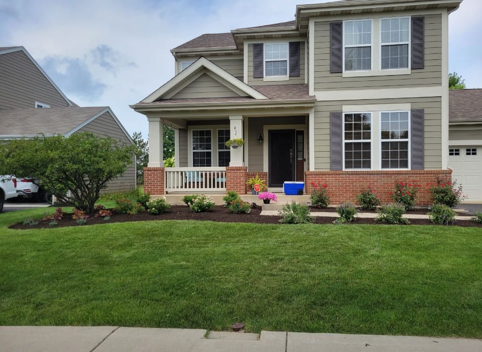 Two-story house with green siding, brick accents, and manicured lawn. Landscaping with flowers and shrubs.